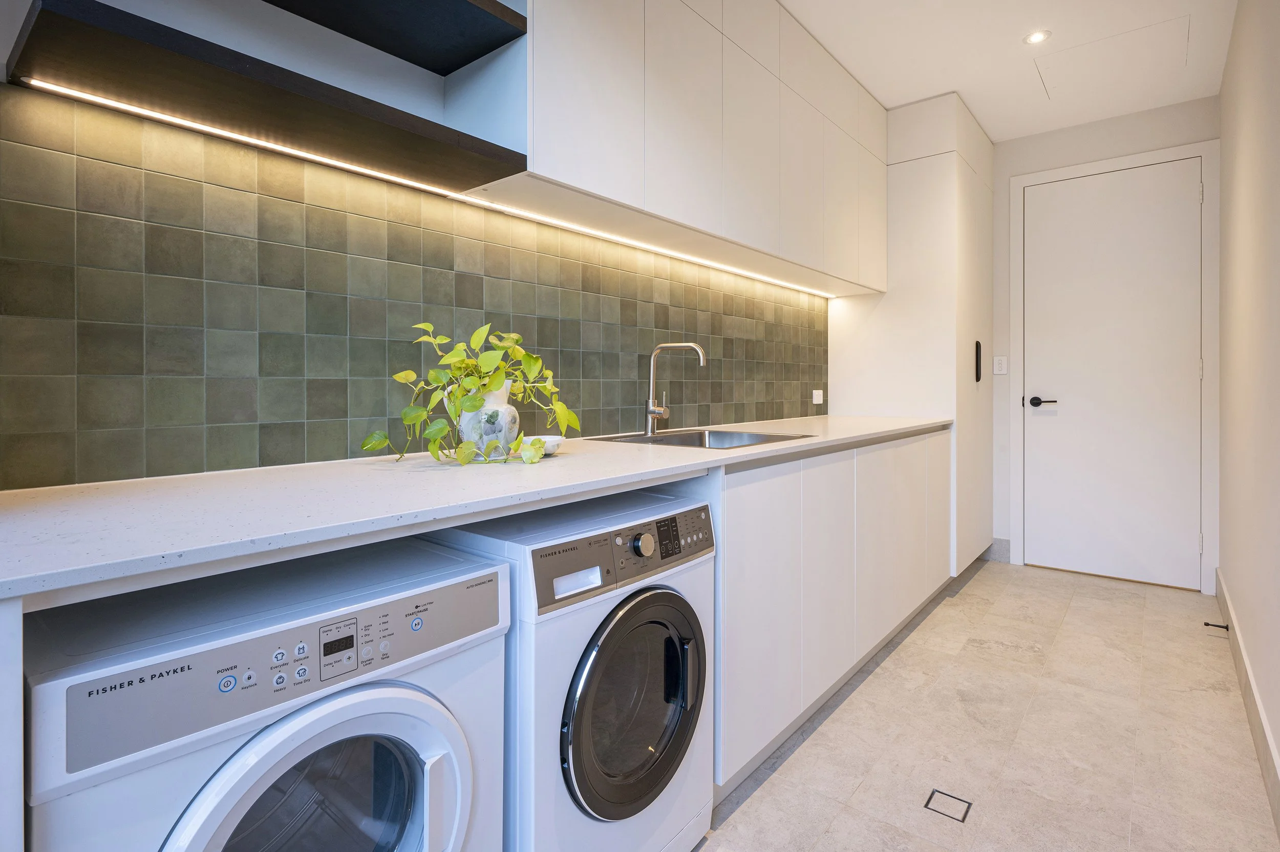 A modern laundry room with white cabinetry, a beige countertop, and a green tiled backsplash. There are two front-loading washing machines stacked beneath the countertop, a sink with a stainless steel faucet, a small plant in a white vase on the coun