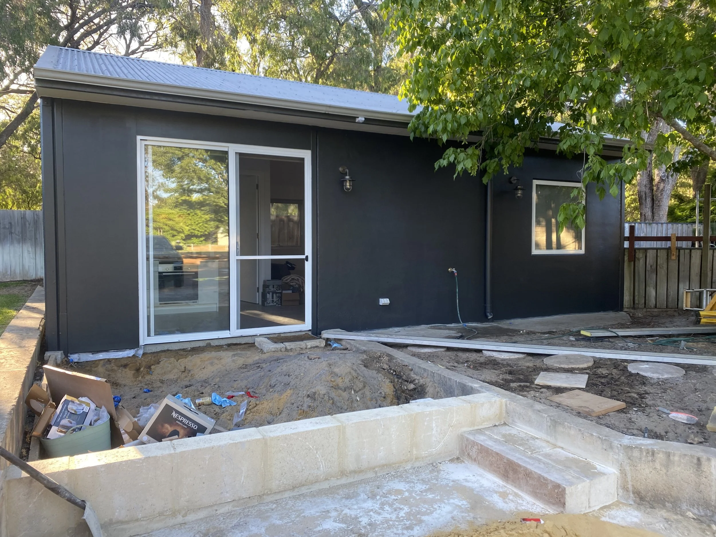 Photo of a backyard patio construction site with a gray house wall, sliding glass door, large window, and a pile of construction debris and tools in the foreground, surrounded by mature trees and a wooden fence.