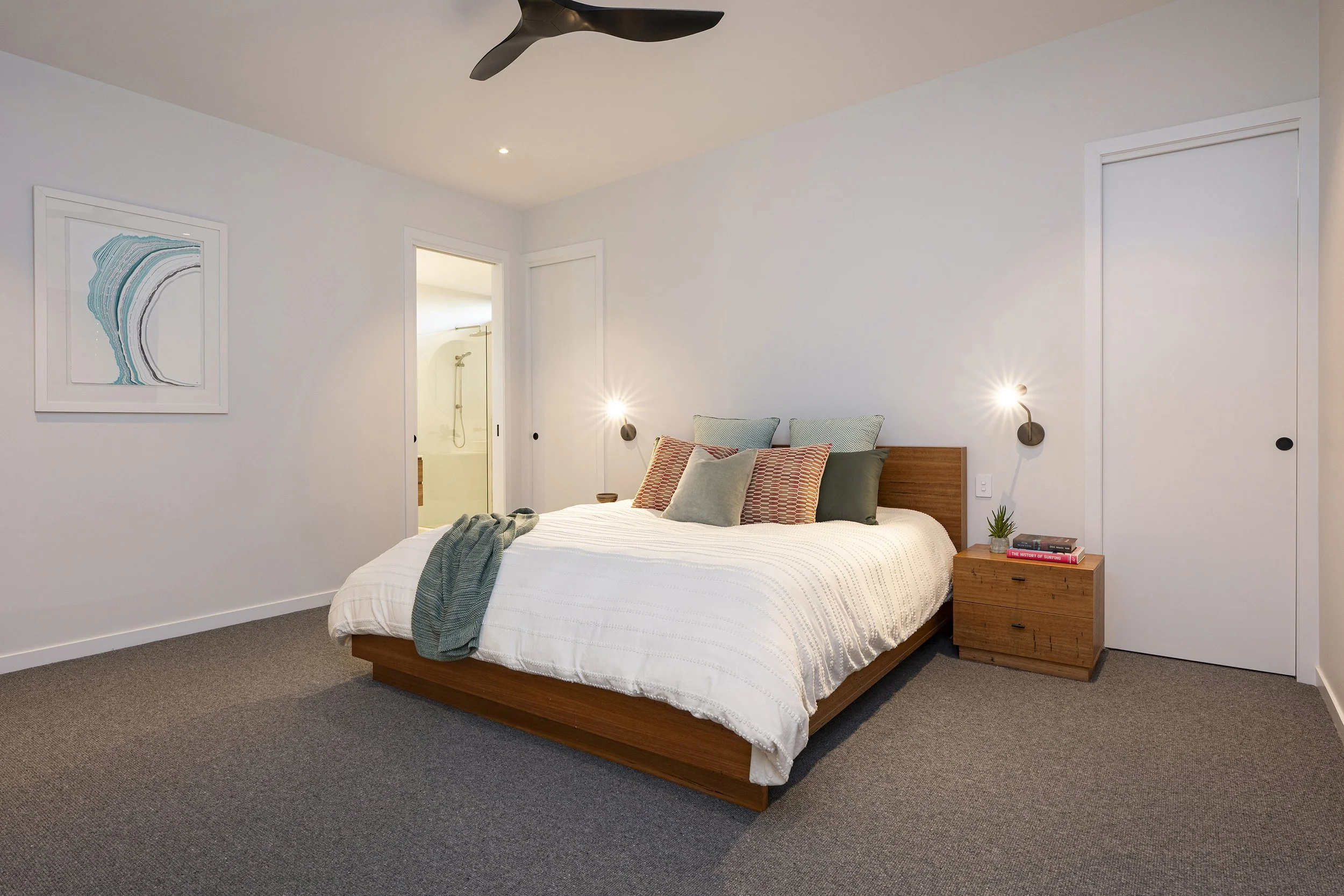 A bedroom with a wooden bed frame, white bedding, and a variety of pillows. There is a wooden nightstand with books and a small plant. The wall has a framed abstract artwork, and there are two wall-mounted reading lights on either side of the bed. Th