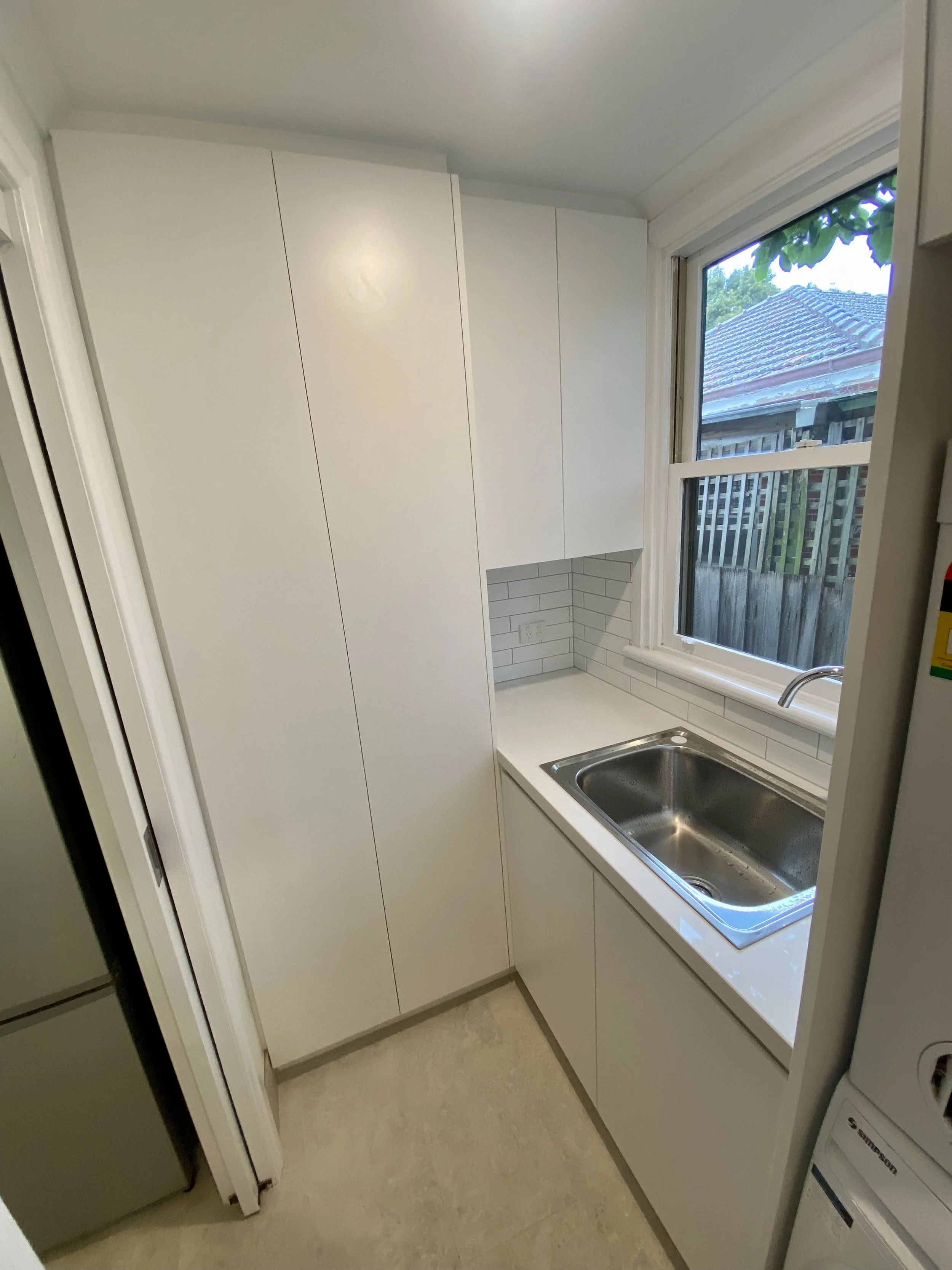 A small kitchen with white cabinets and a stainless steel sink under a window showing a backyard with a roof and trees.