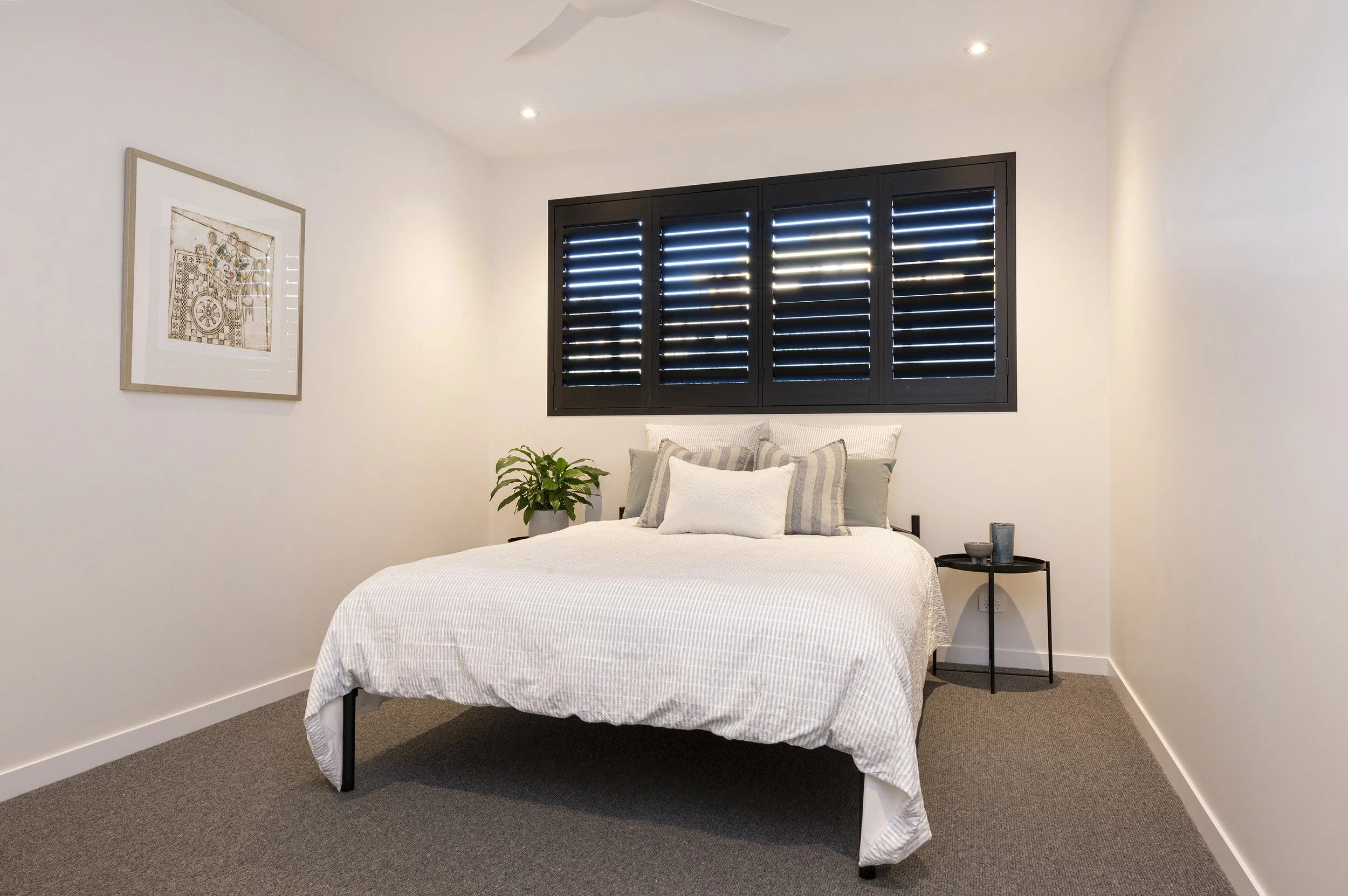 A bedroom with white walls, a bed with white and striped pillows, a black window with shutters, a framed artwork on the wall, a small black side table with decorative items, a green plant, and ceiling lights.