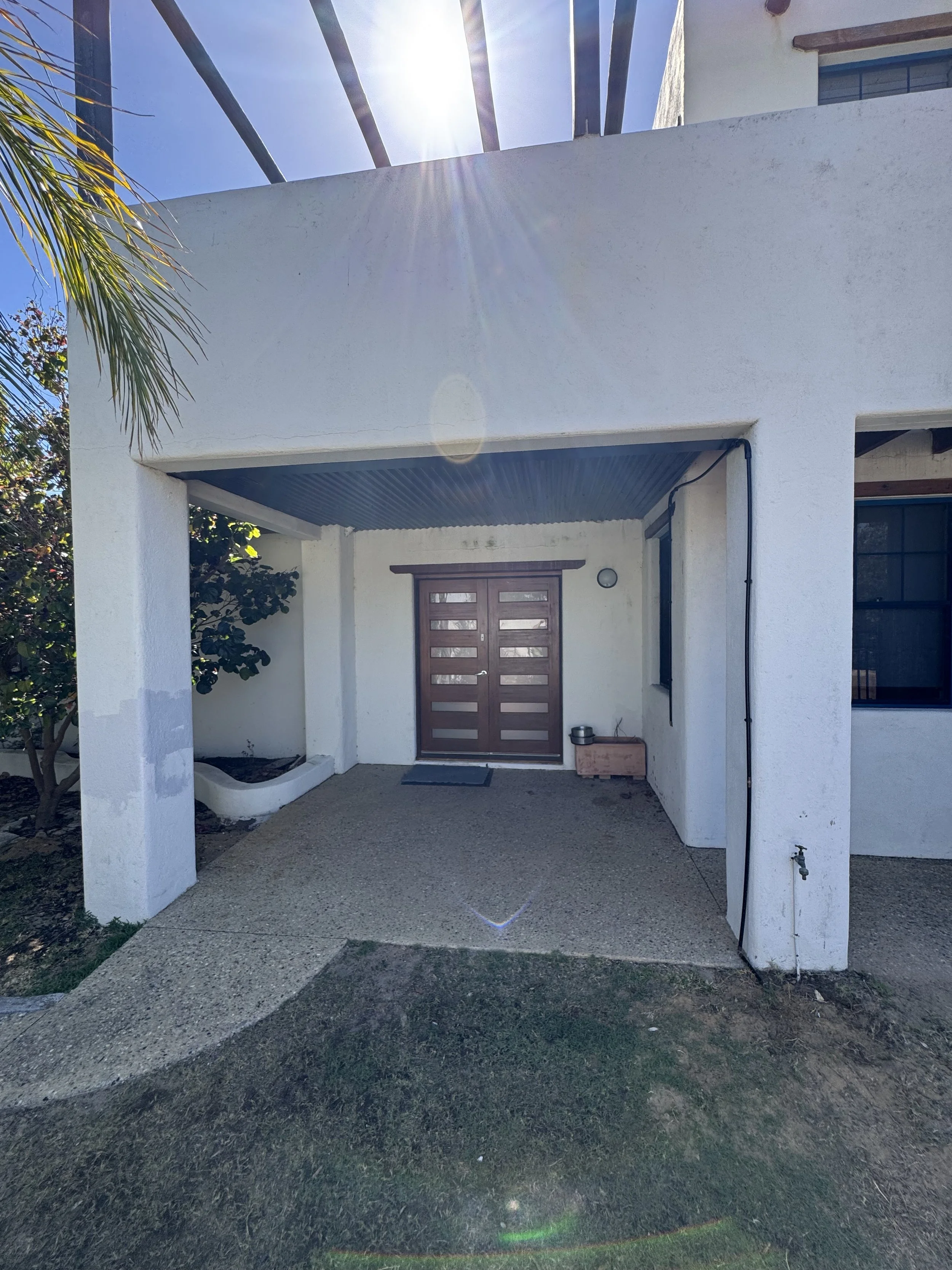 Front view of a white modern house with a wooden door, a small porch, and a concrete pathway. Sunlight shines brightly from above, casting shadows on the ground. There is a palm tree partially visible on the left side.