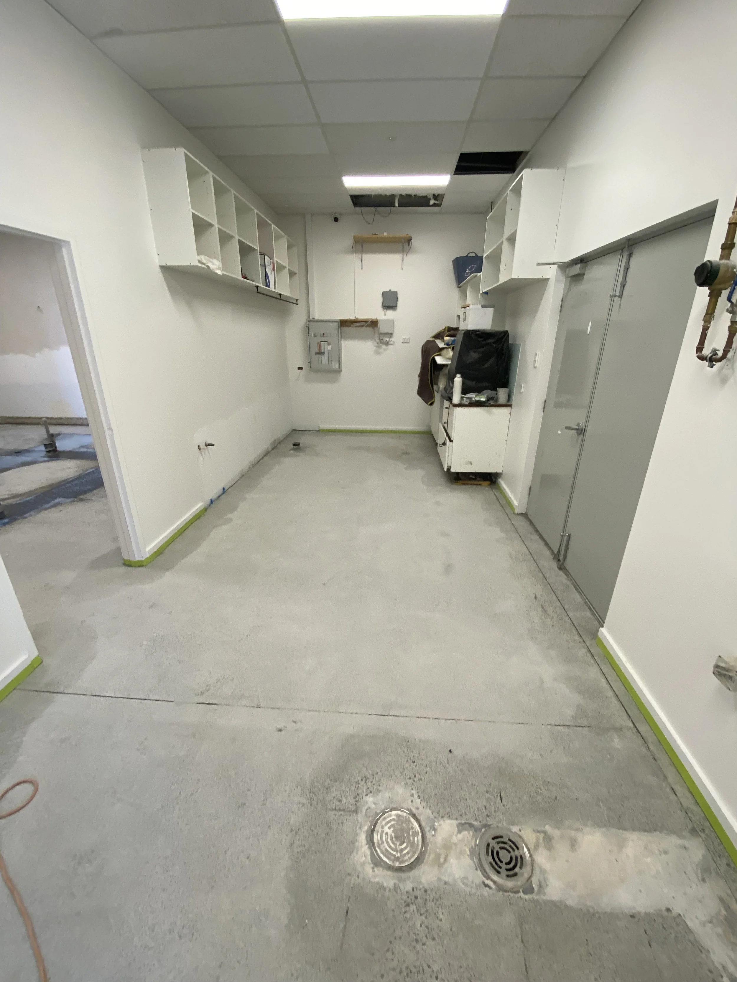 A room under construction with unfinished flooring, white walls with green tape on the baseboards, a ceiling with missing and new tiles, and various items including a chair, boxes, and open shelving.