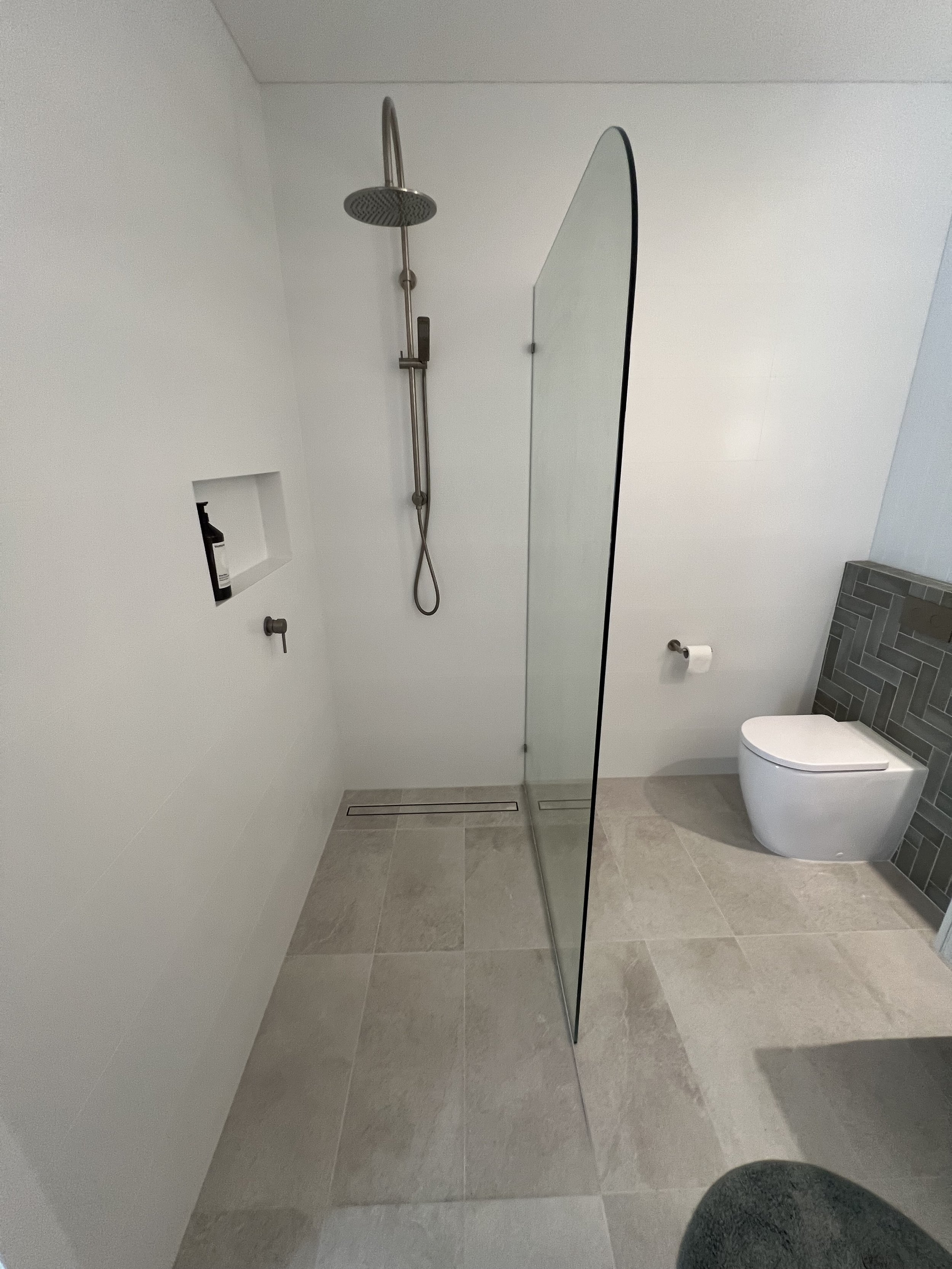 Modern bathroom with white walls, gray tile flooring, a walk-in shower with a glass partition, wall-mounted showerhead and faucet, built-in wall niche with toiletries, and a wall-mounted toilet with a toilet paper holder.