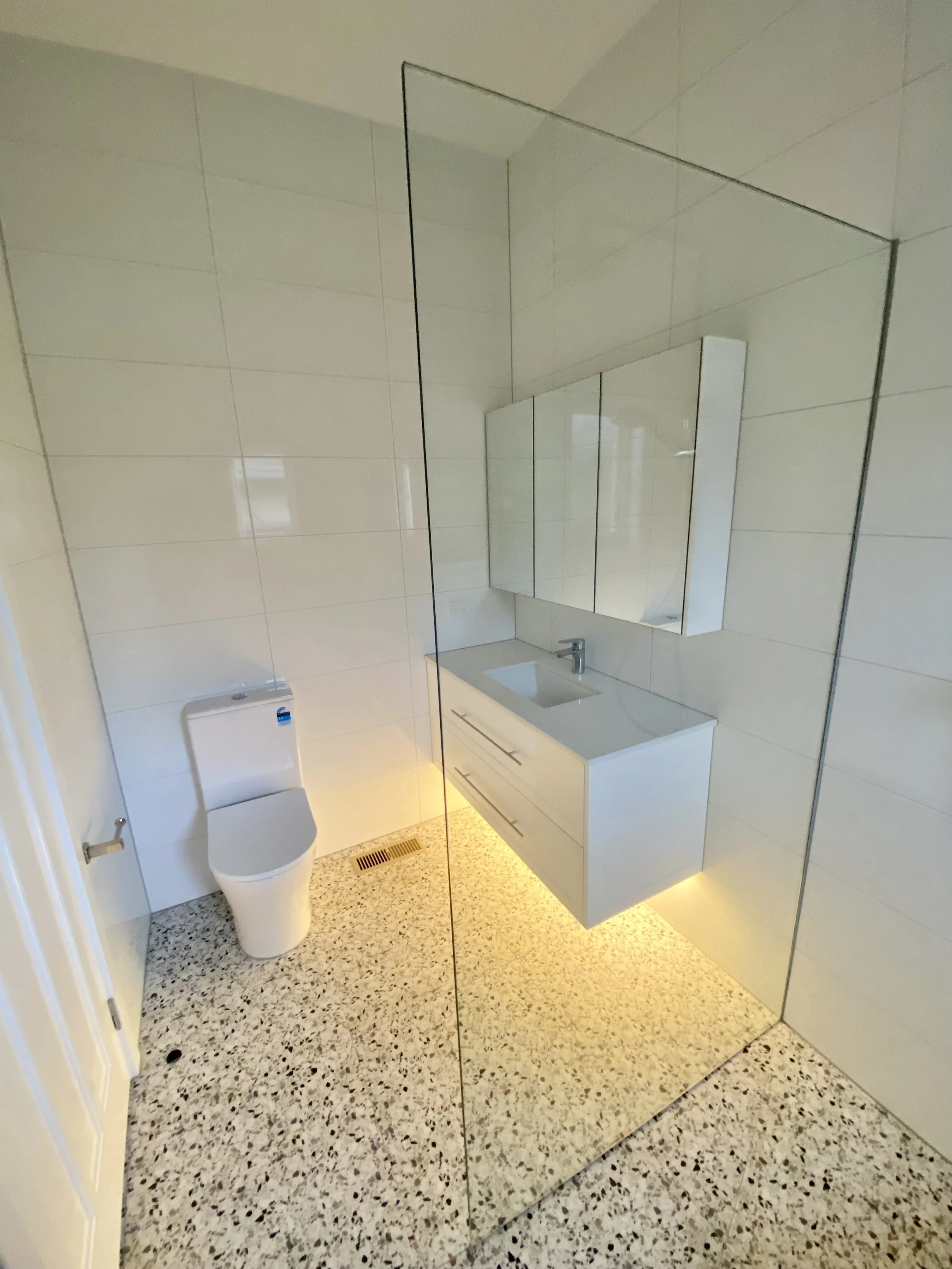 A modern bathroom featuring a white toilet, a floating white vanity with a sink, a mirror, tiled walls, terrazzo flooring, and a floor vent.