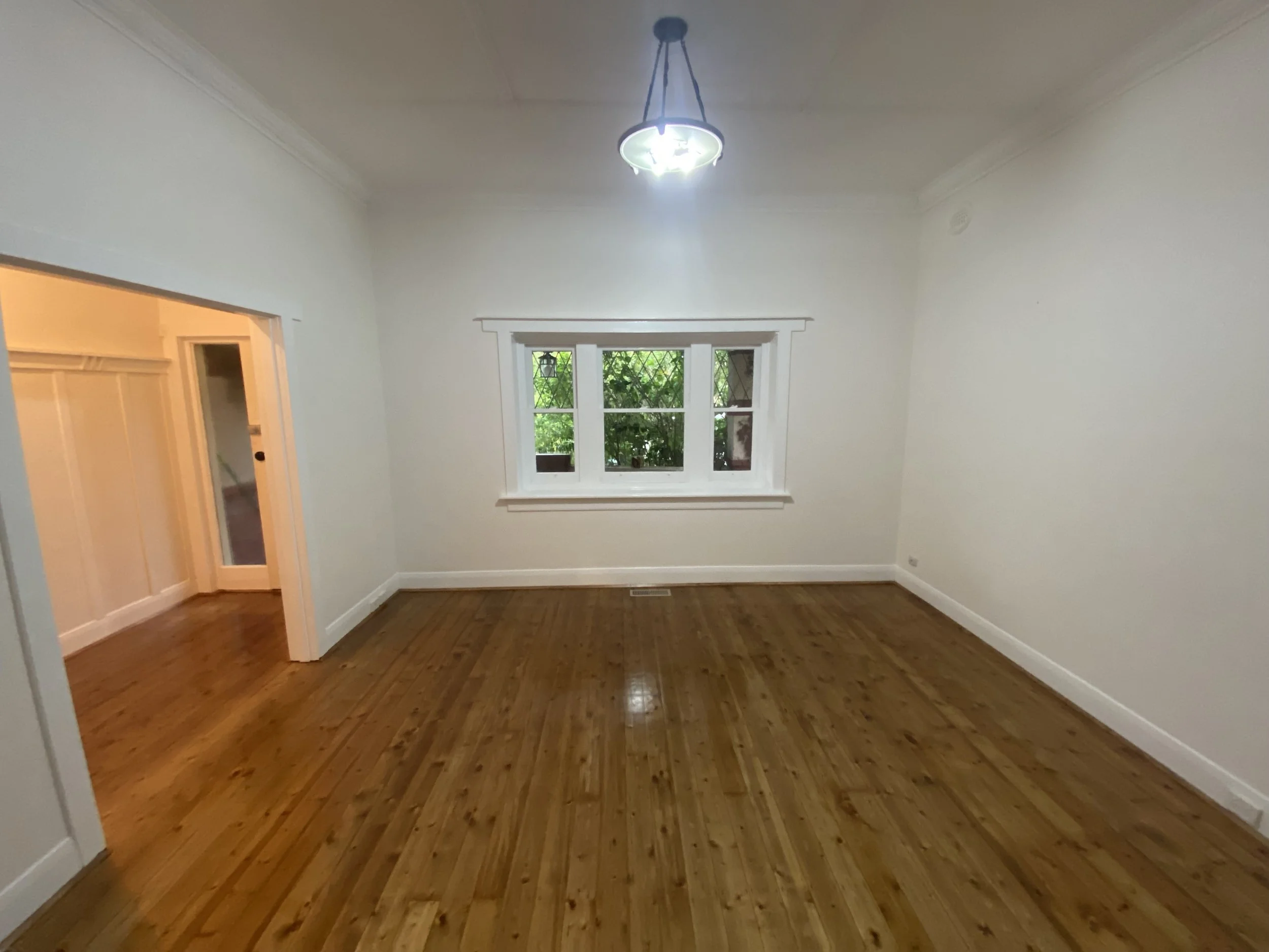 Empty room with hardwood floors, white walls, a window with greenery outside, and a ceiling light fixture.