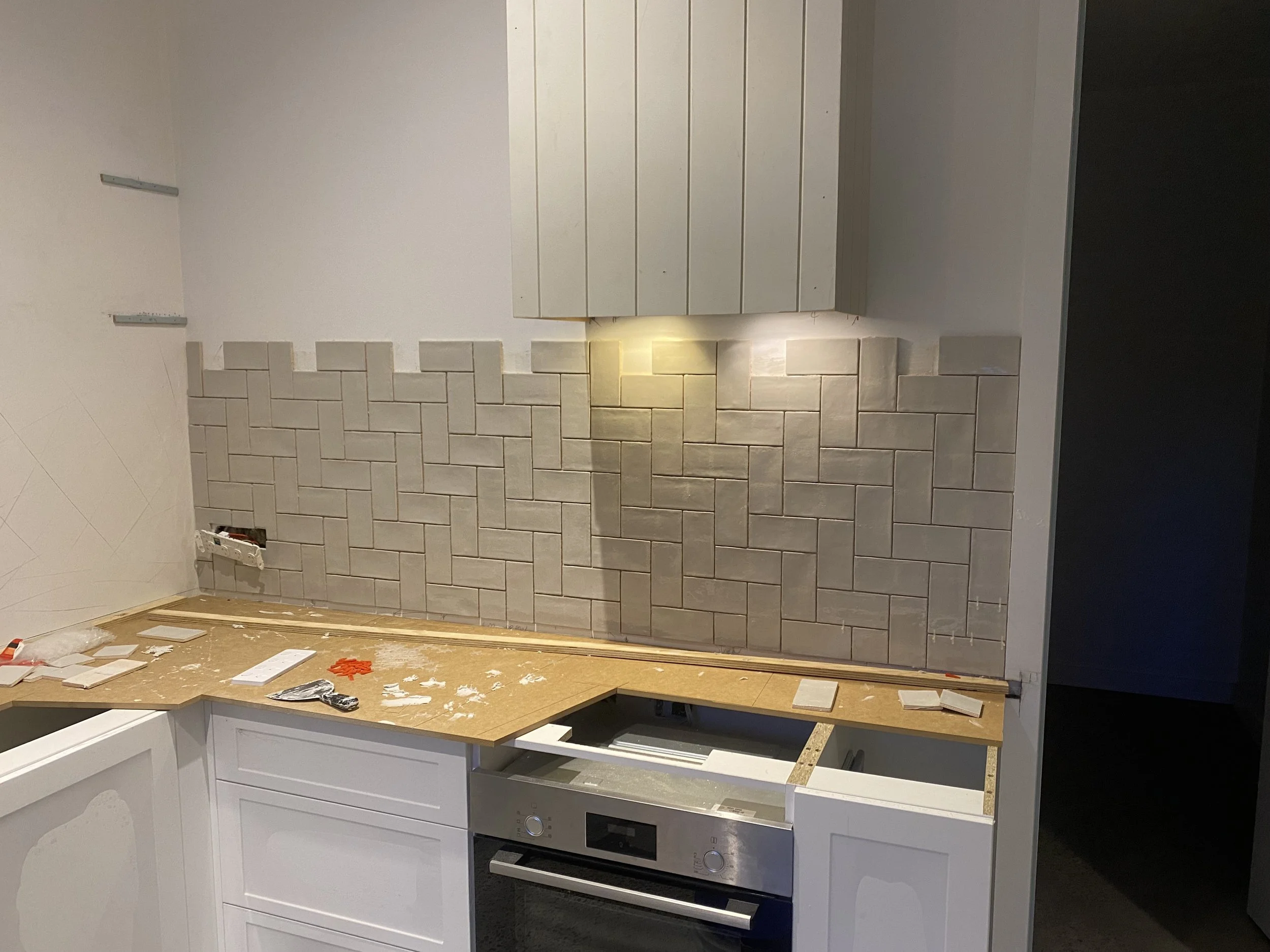 Kitchen wall with beige brick tile backsplash, partially installed white cabinetry, and a built-in oven. There are tools and tiles scattered on the countertop.