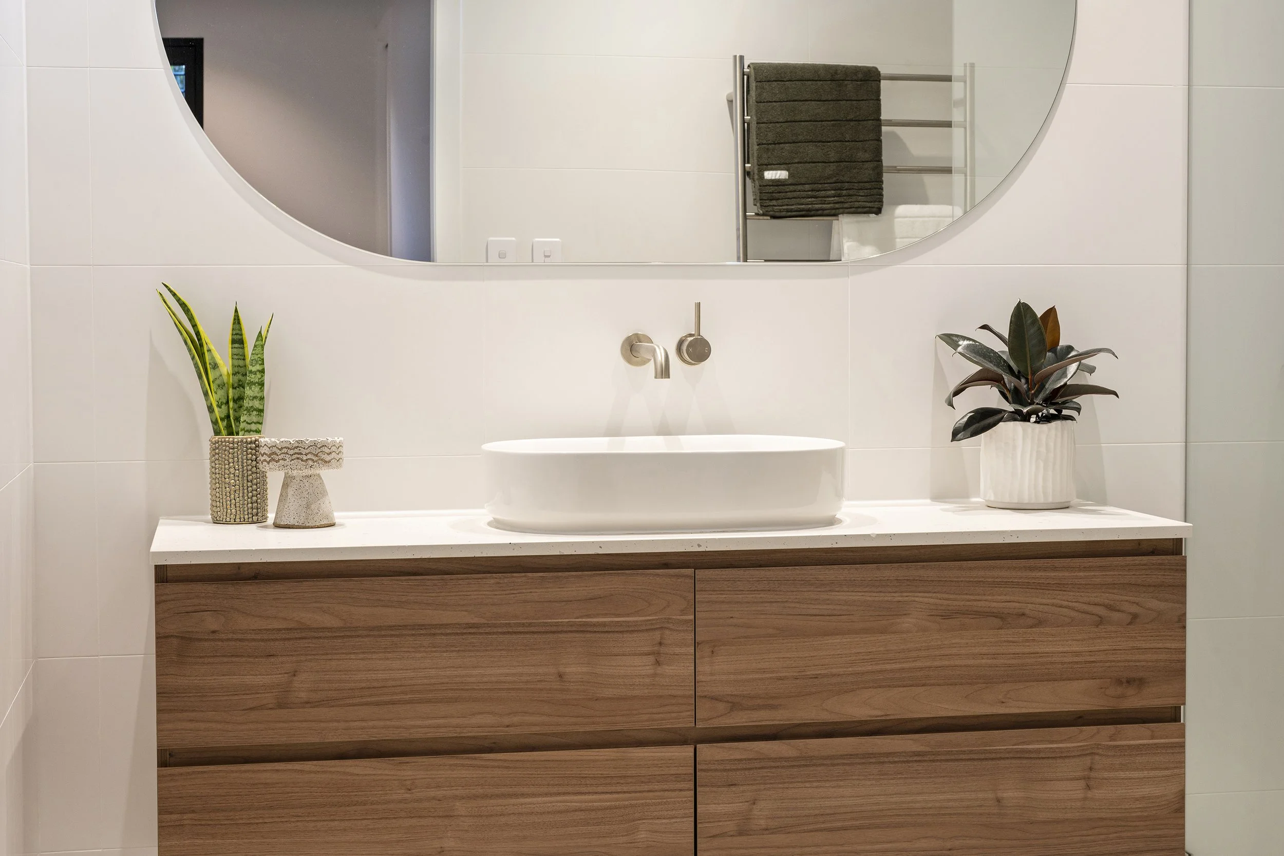 A bathroom vanity with a white vessel sink, two potted plants in decorative pots on either side, a large mirror above, and a towel rack with folded dark gray towels in the background.
