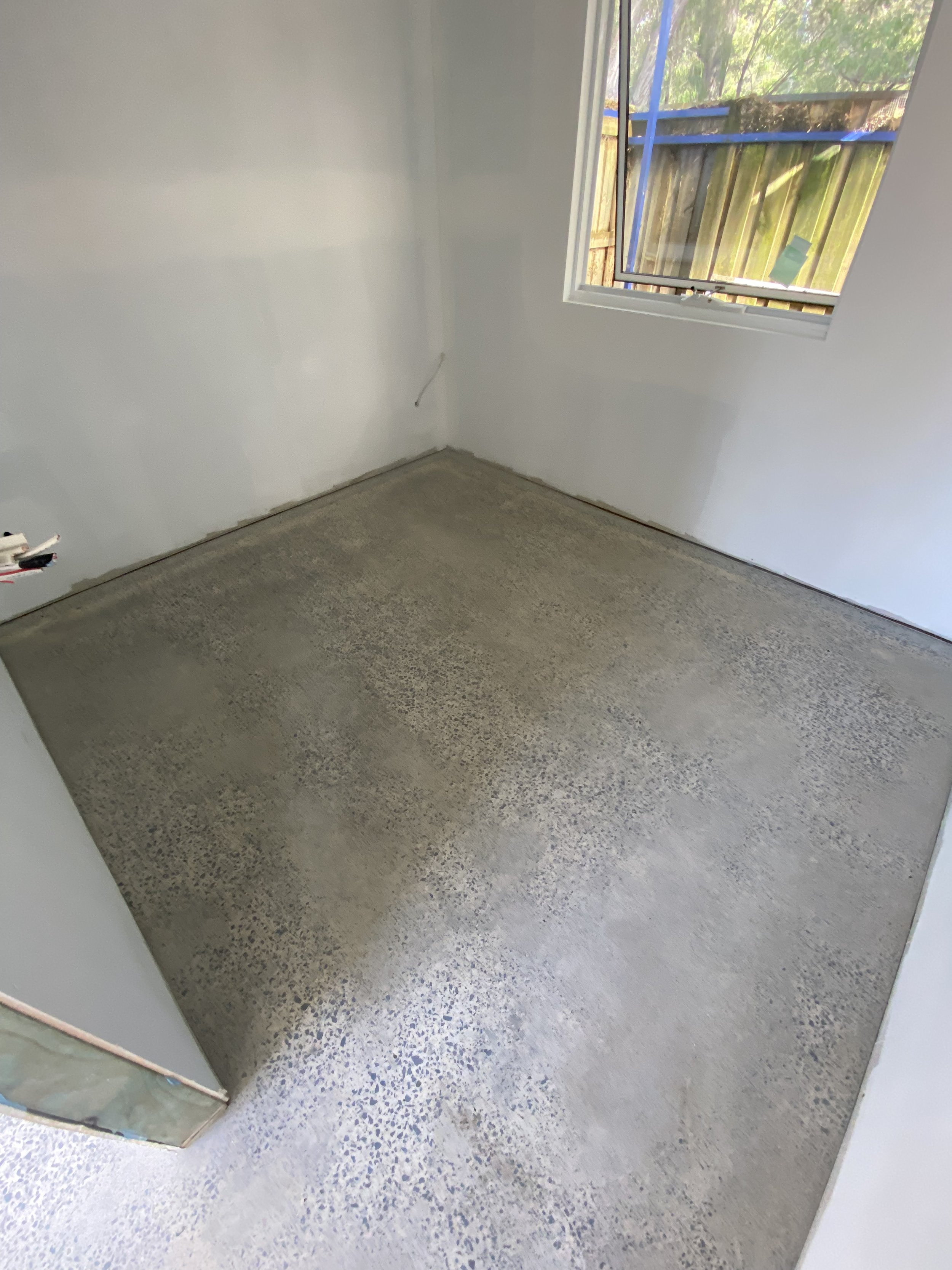 Empty room with unfinished flooring, white walls, and a large window showing a wooden fence and trees outside.