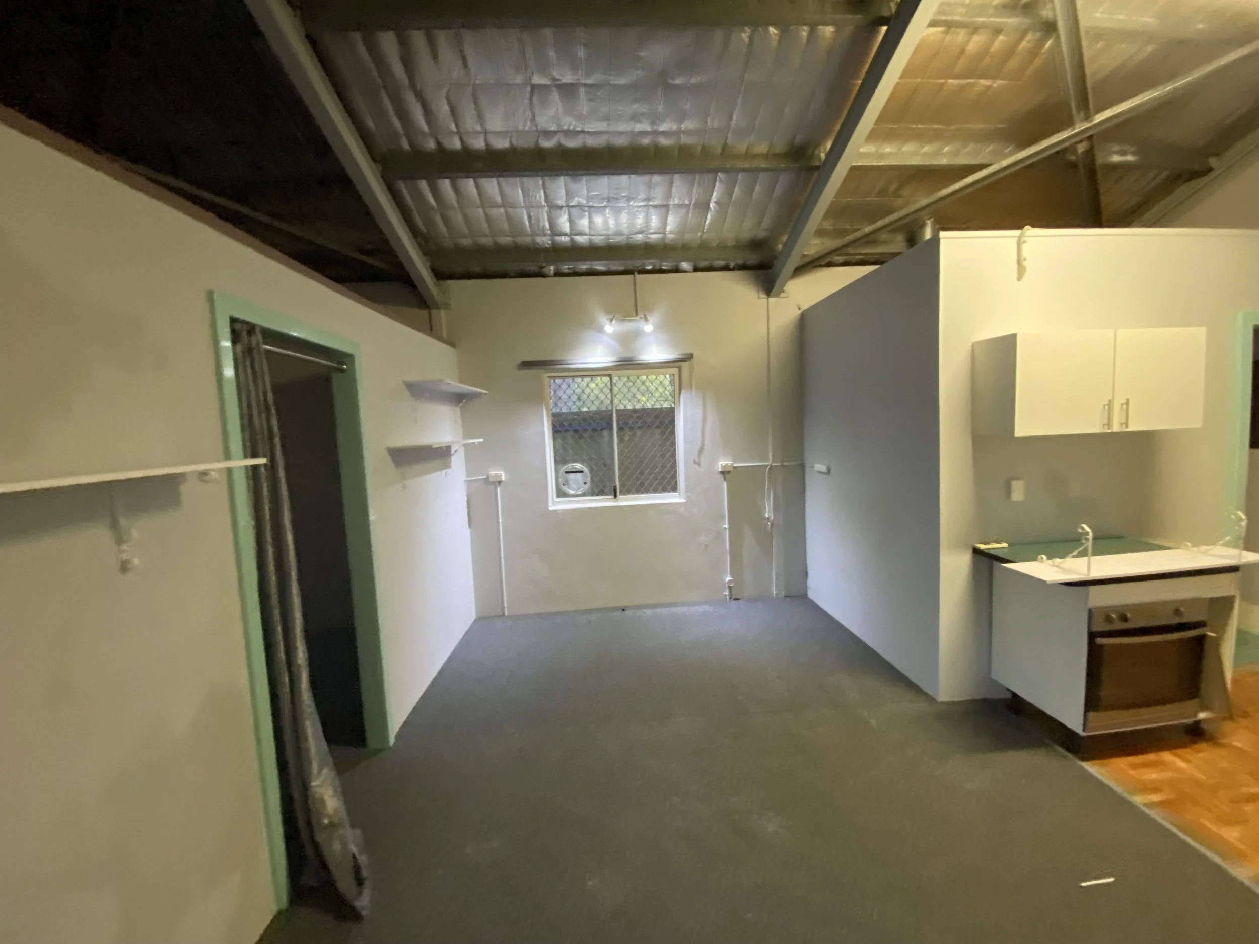 Empty room with gray carpet, beige walls with green trim, ceiling with metal beams and insulation, small window with security screen, and partial view of kitchen with white cabinets.