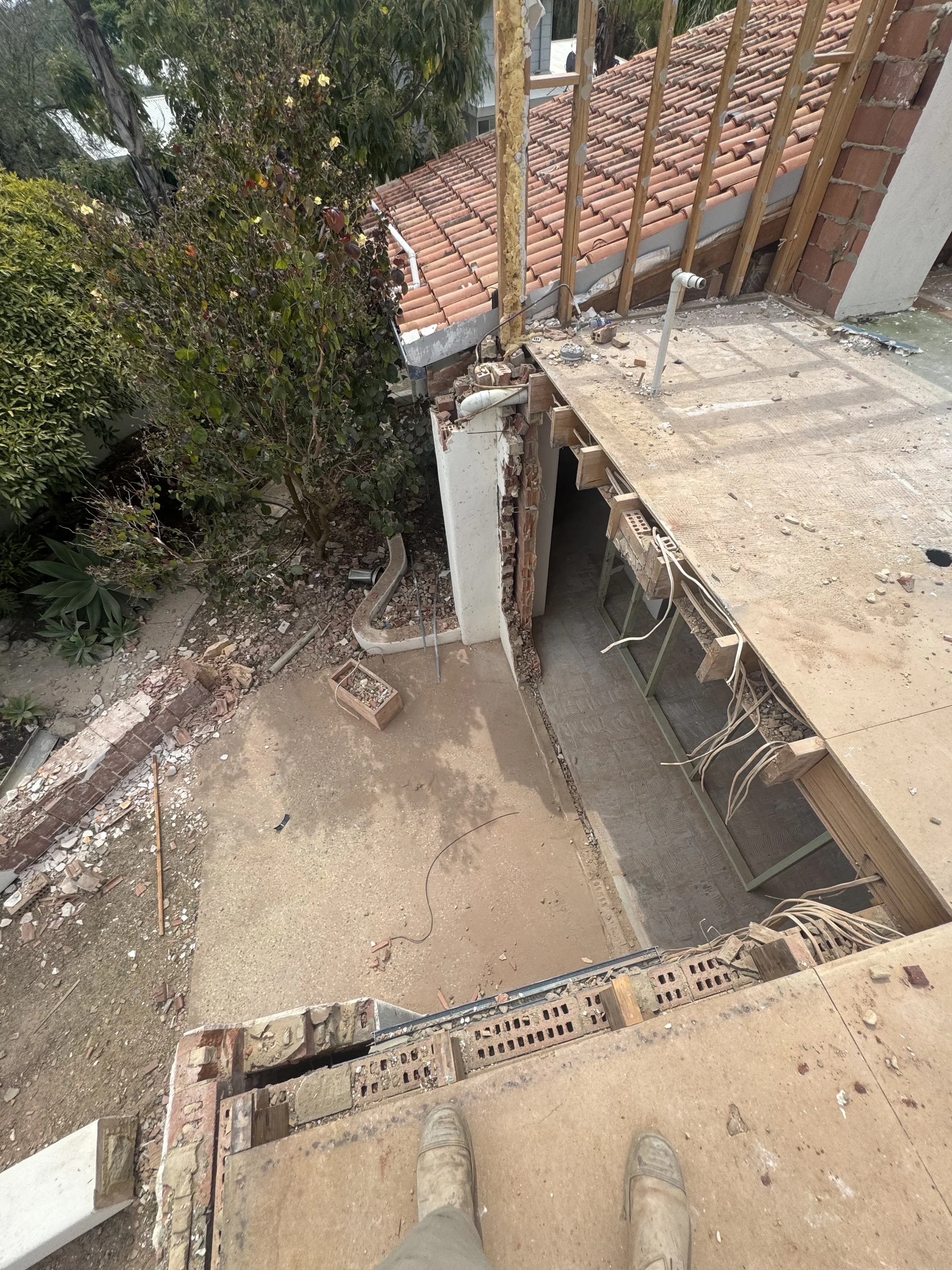 View from a construction site looking down at a dirt floor with partially built brick wall, exposed wooden framework, measuring tools, and construction debris. The photo shows the edge of a building with a tiled roof and lush green trees nearby.