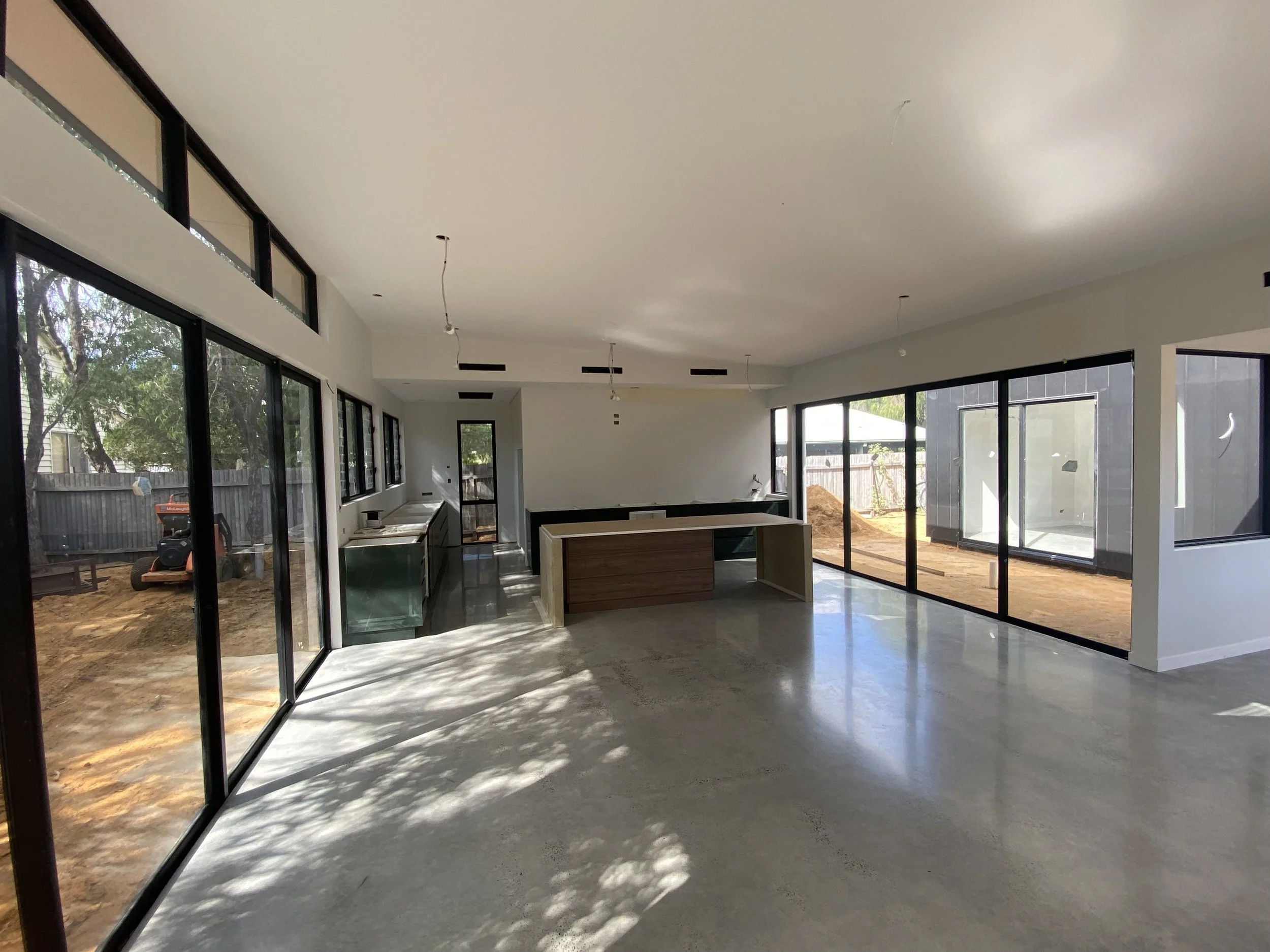 Interior view of a house under construction with large glass windows and sliding doors, unfinished floors, and visible ceiling wiring.