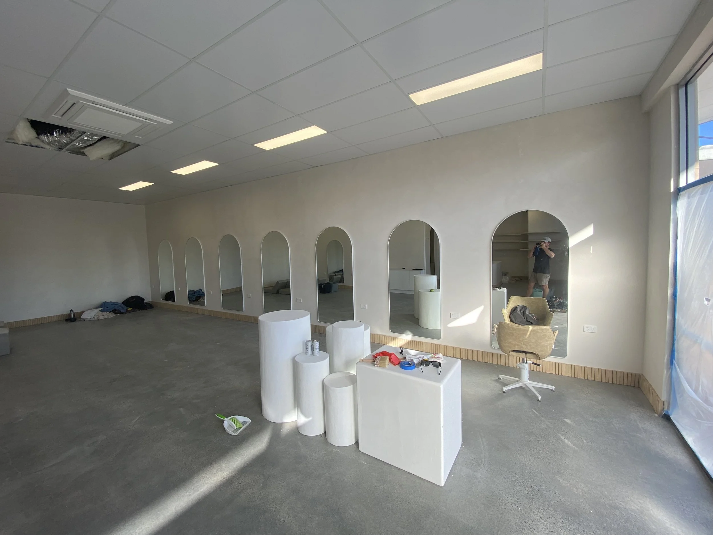 Empty commercial space with a partially installed ceiling, scattered furniture, and construction tools, including a person working near the wall.