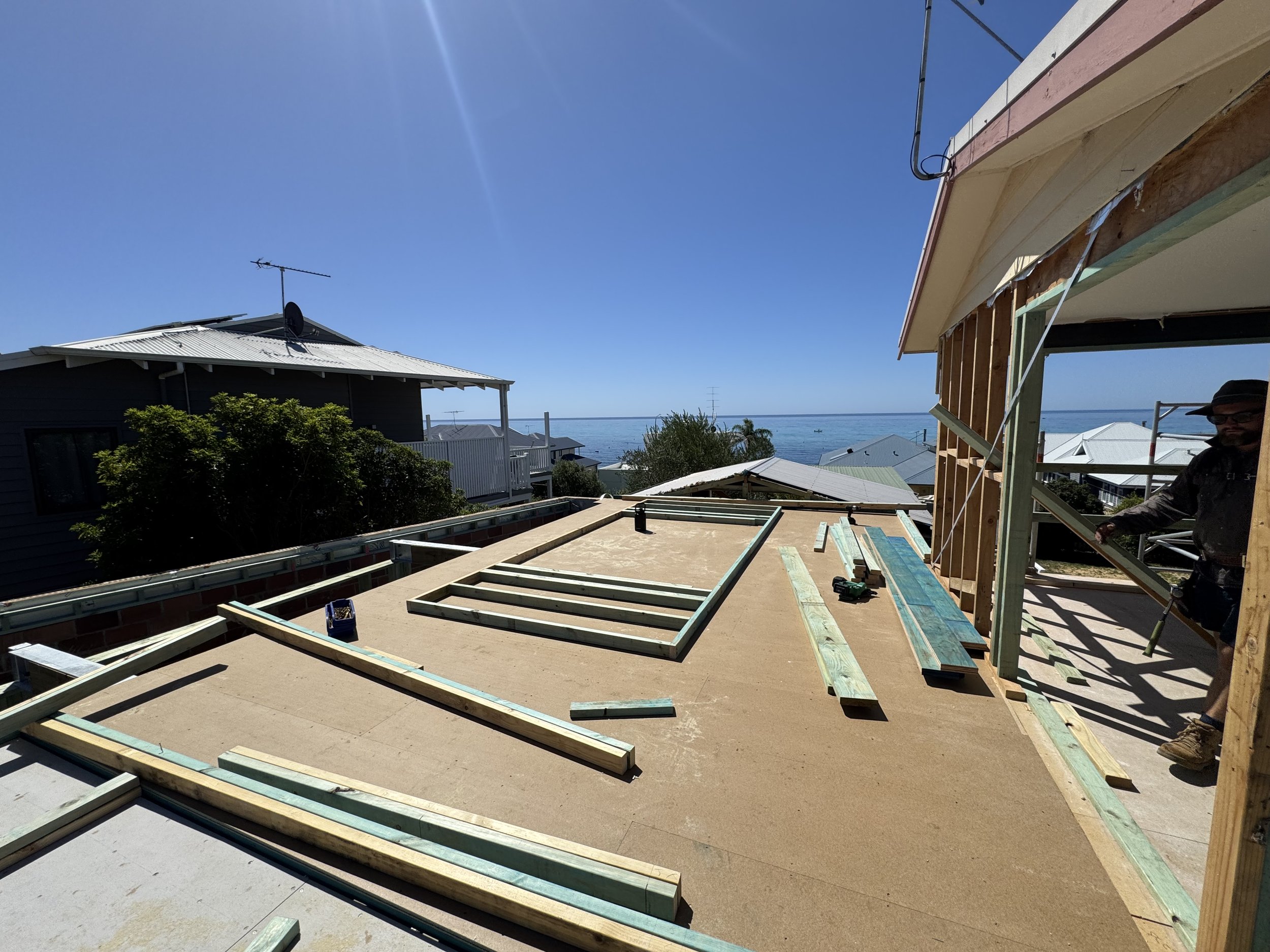 Construction site of a house with a view of the ocean in the background. The house structure is in the framing stage with wooden beams and plywood flooring, with roofing work ongoing.