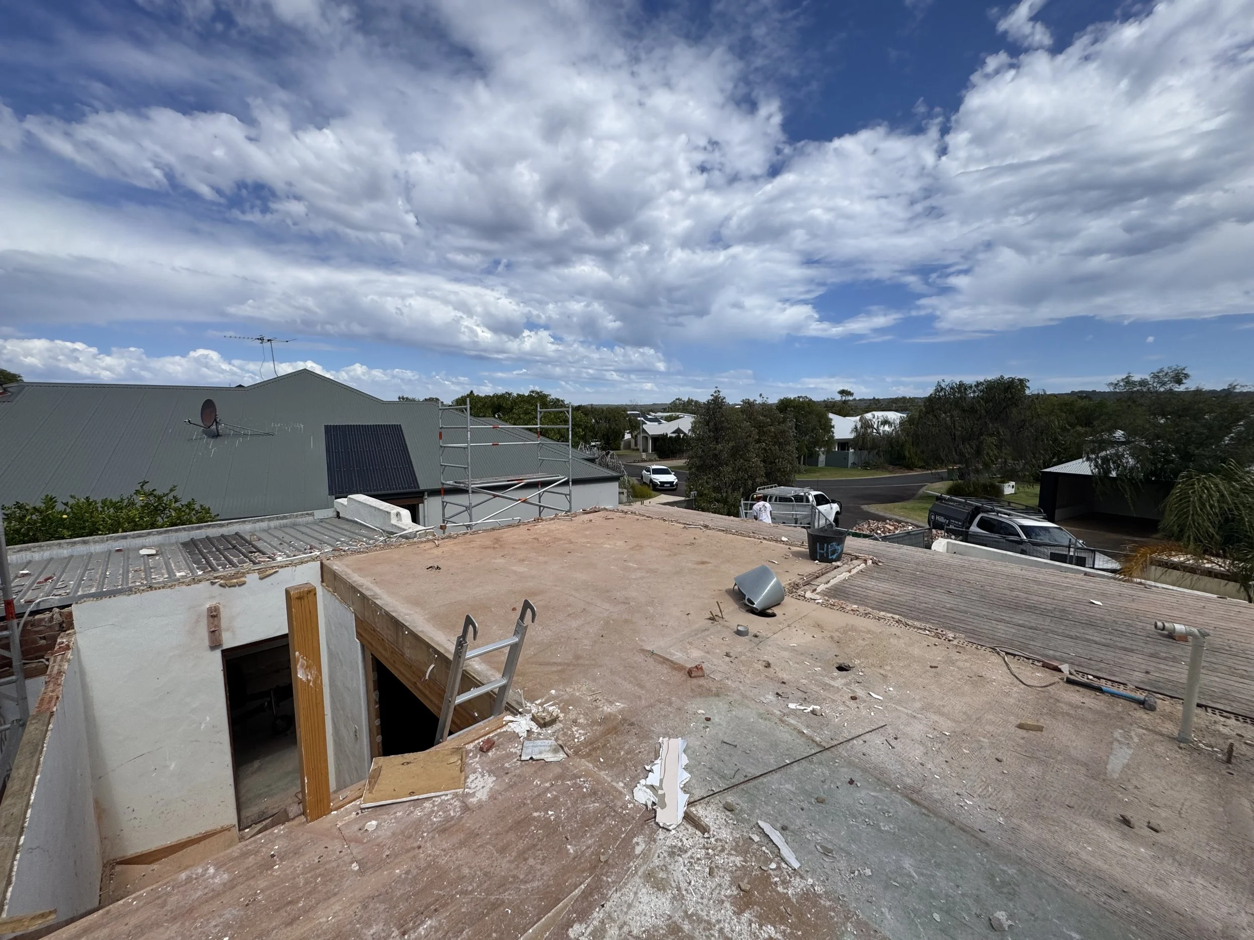 View from a rooftop under construction or renovation showing a partly finished wooden deck, construction debris, and nearby houses with trees under a blue sky with scattered clouds.