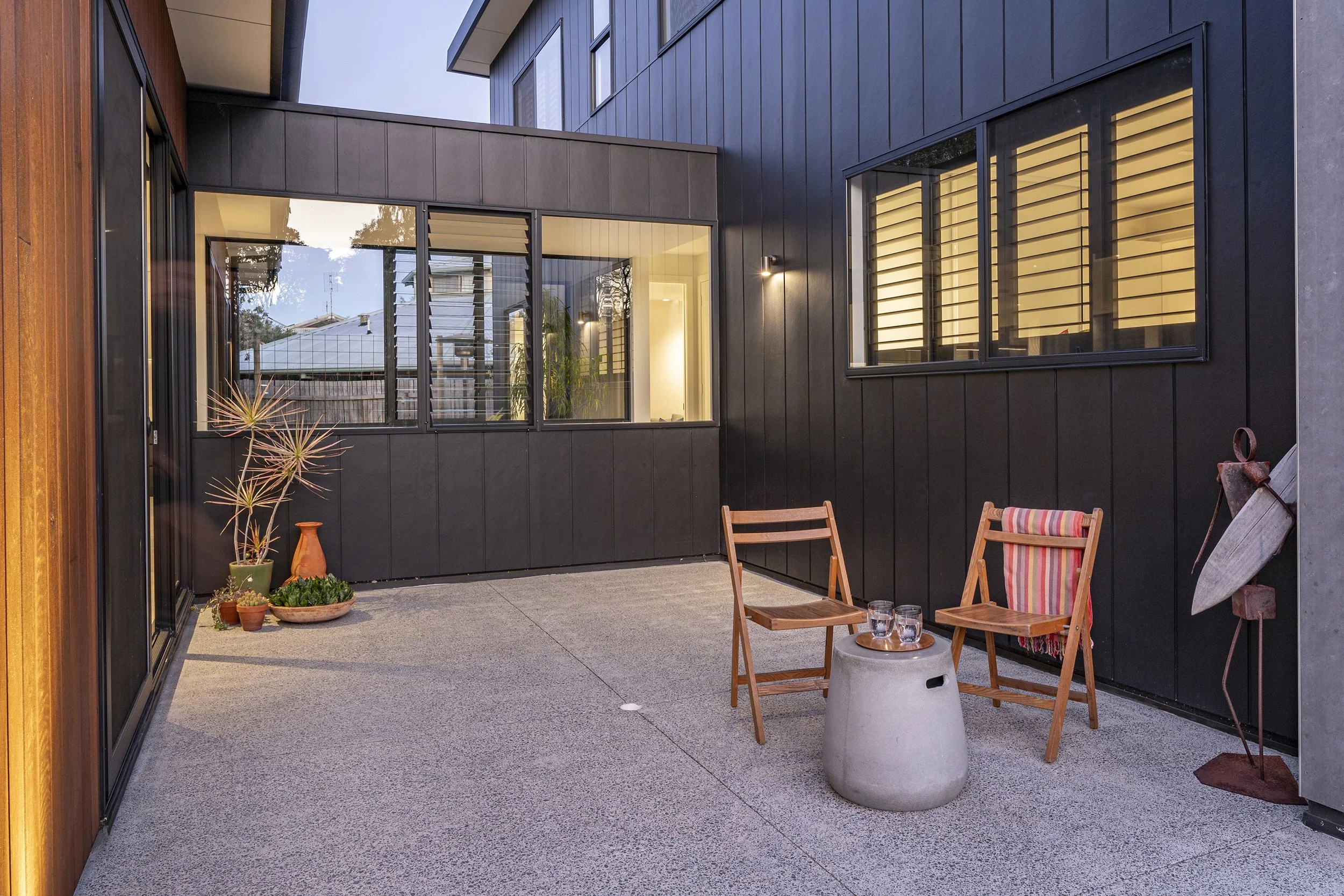 A modern outdoor patio with black and wooden siding, large windows with blinds, a small round concrete table, wooden chairs, potted plants, and a glass of water.