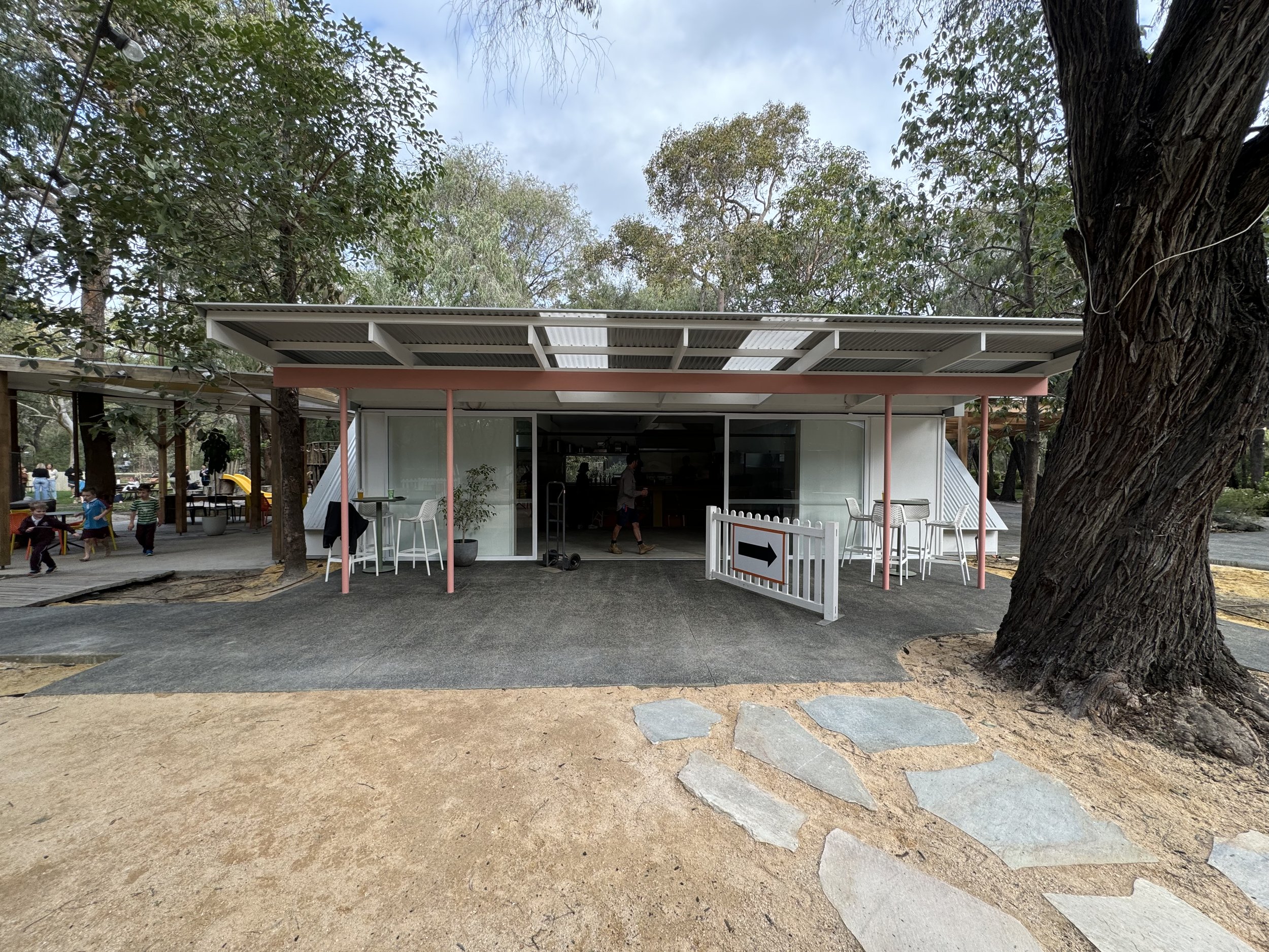 Outdoor scene at a park with children playing, a building with a patio, and large trees surrounding the area.