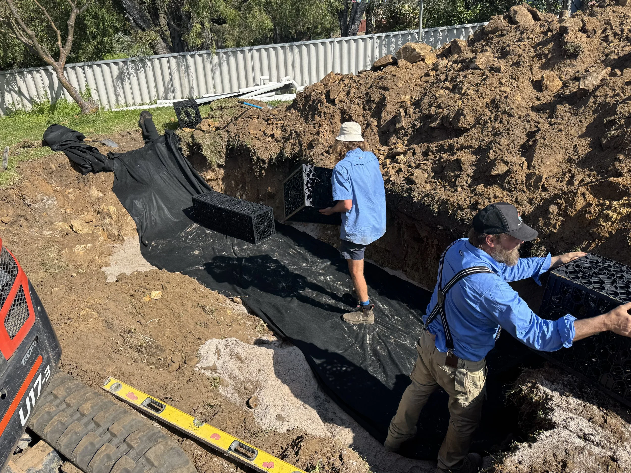 People working on a construction or excavation site in a backyard, digging a trench, with piles of dirt, tools, and safety gear visible.