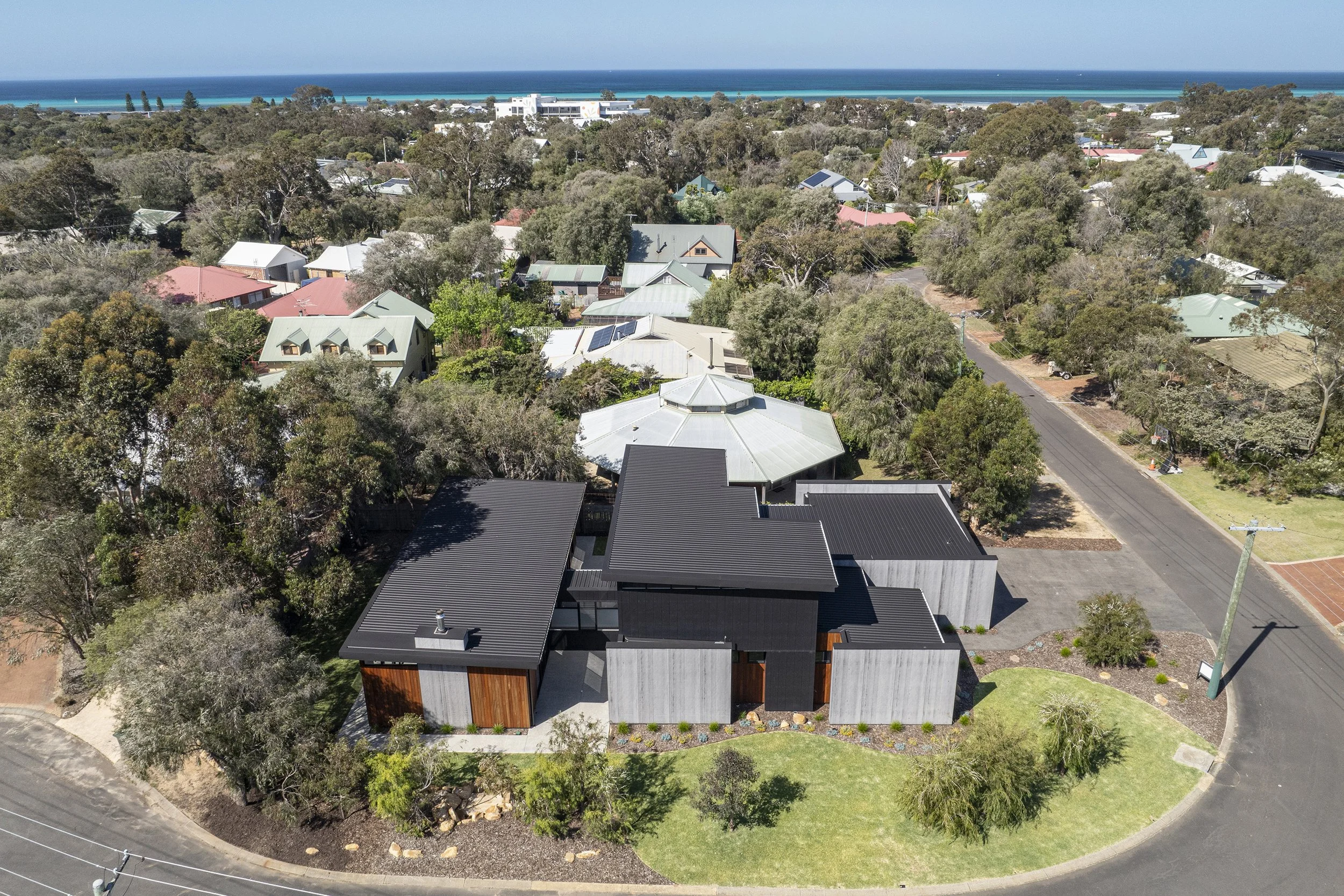 Aerial view of a residential neighborhood near the coast with houses, trees, and a road, with the ocean visible in the background.