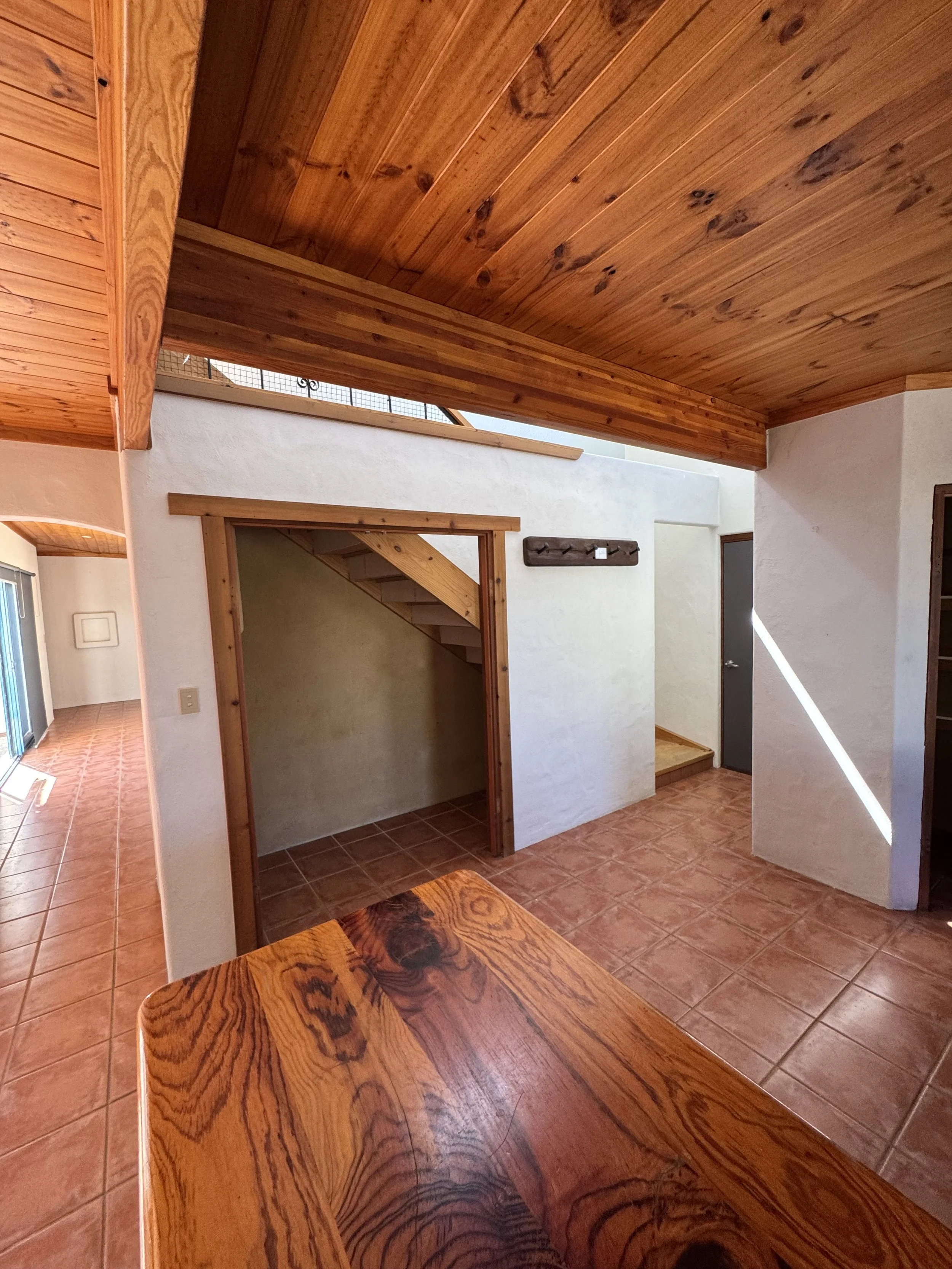 Interior of a house with a wooden ceiling, tiled floor, and white walls. The foreground features a wooden kitchen table with a natural finish, and there is a door framing an open doorway into another room with stairs.
