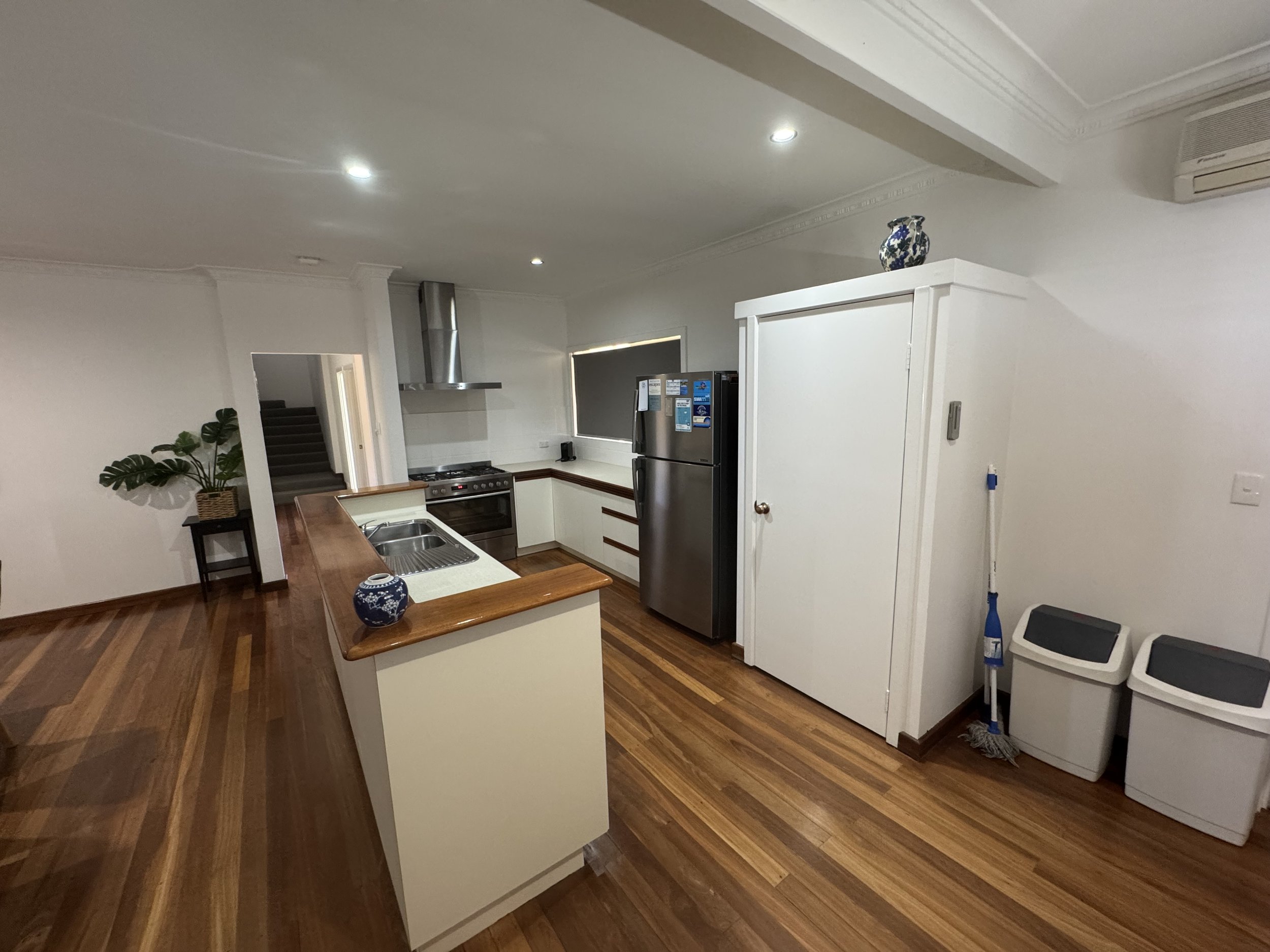 A kitchen with white cabinets, a wooden countertop, a stainless steel refrigerator, a stove, a plant on a small table, and a corner with laundry machines and cleaning supplies.