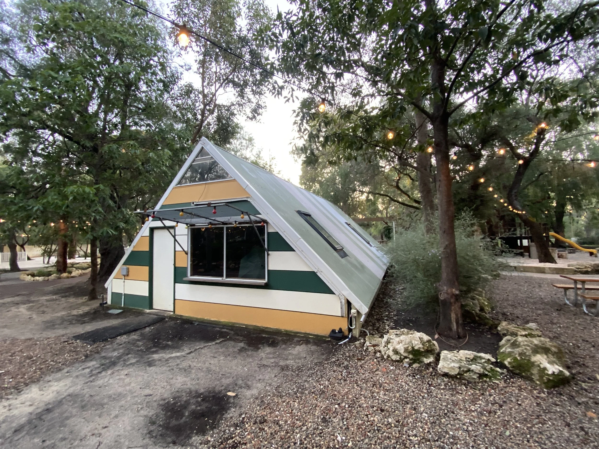 A small green and white house with a sloped metal roof, surrounded by tall trees with string lights hanging from the branches.