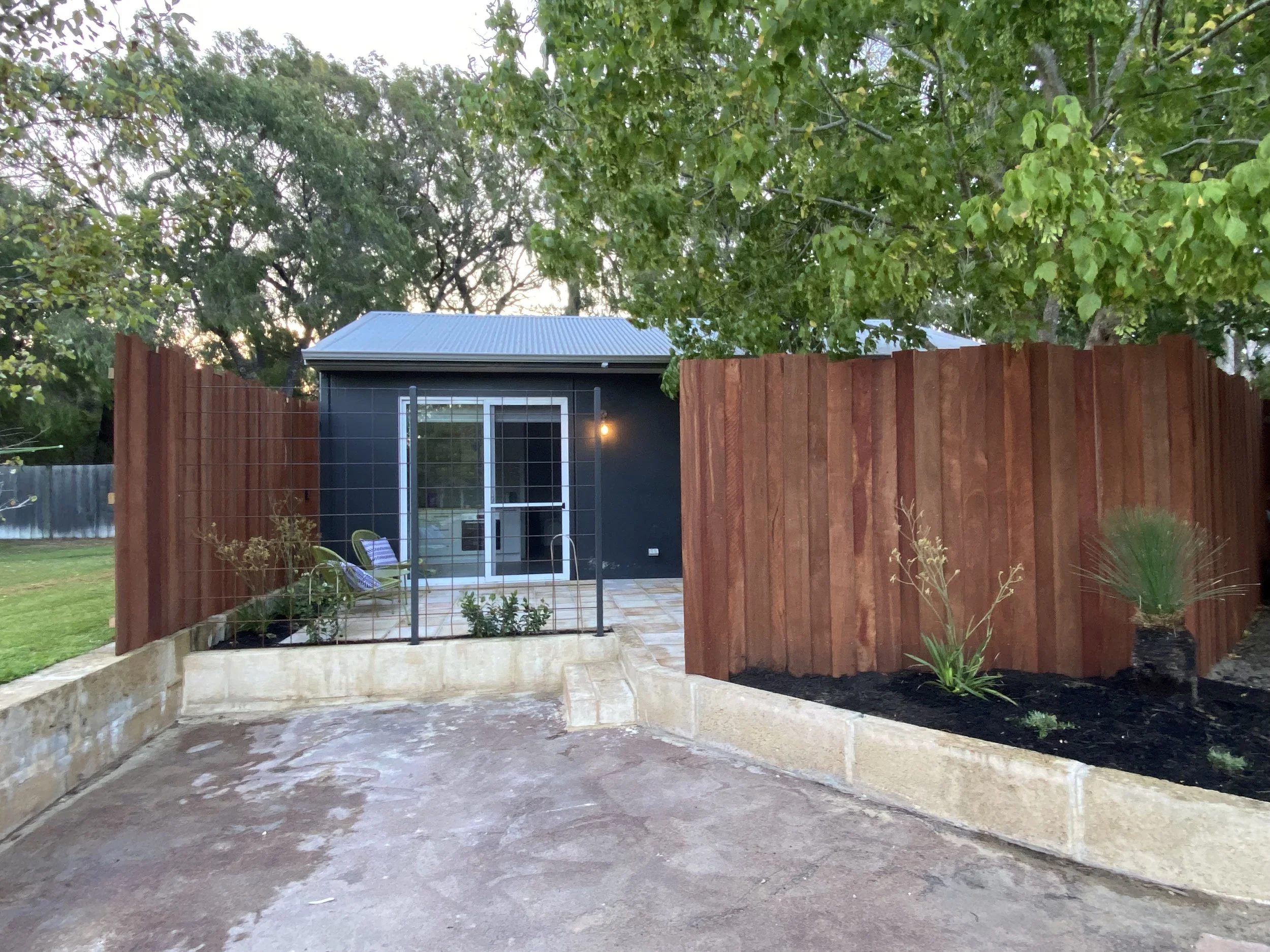 Backyard with a dark-colored shed, a wooden fence, and a patio with potted plants. Large green trees are visible in the background.
