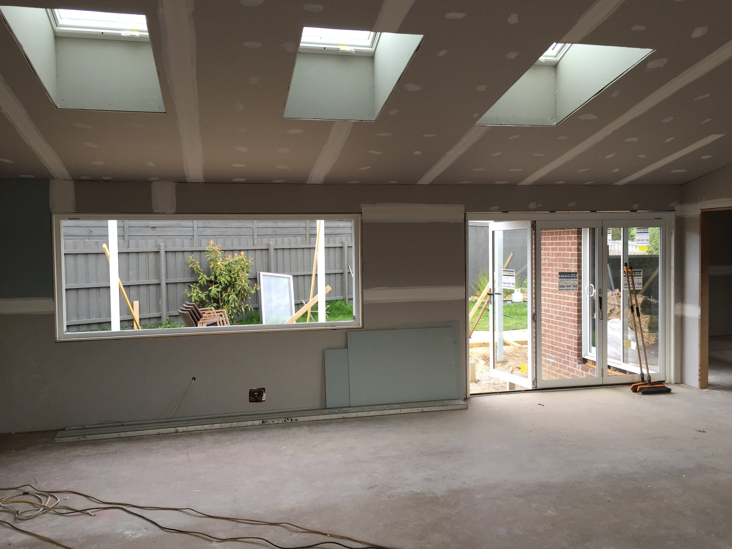 Interior of a house under construction with drywall ceiling, large windows, and sliding glass doors leading outside. Construction materials and tools are visible.