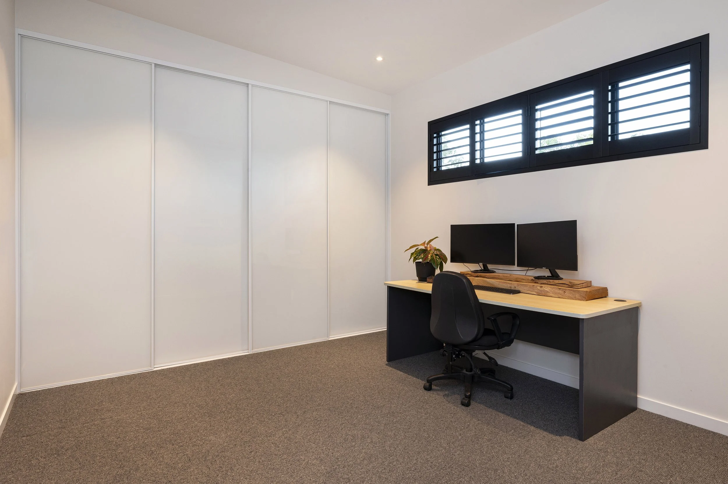 An office corner with a white wall, black window shutters, a desk with two monitors, a plant, and a black office chair.