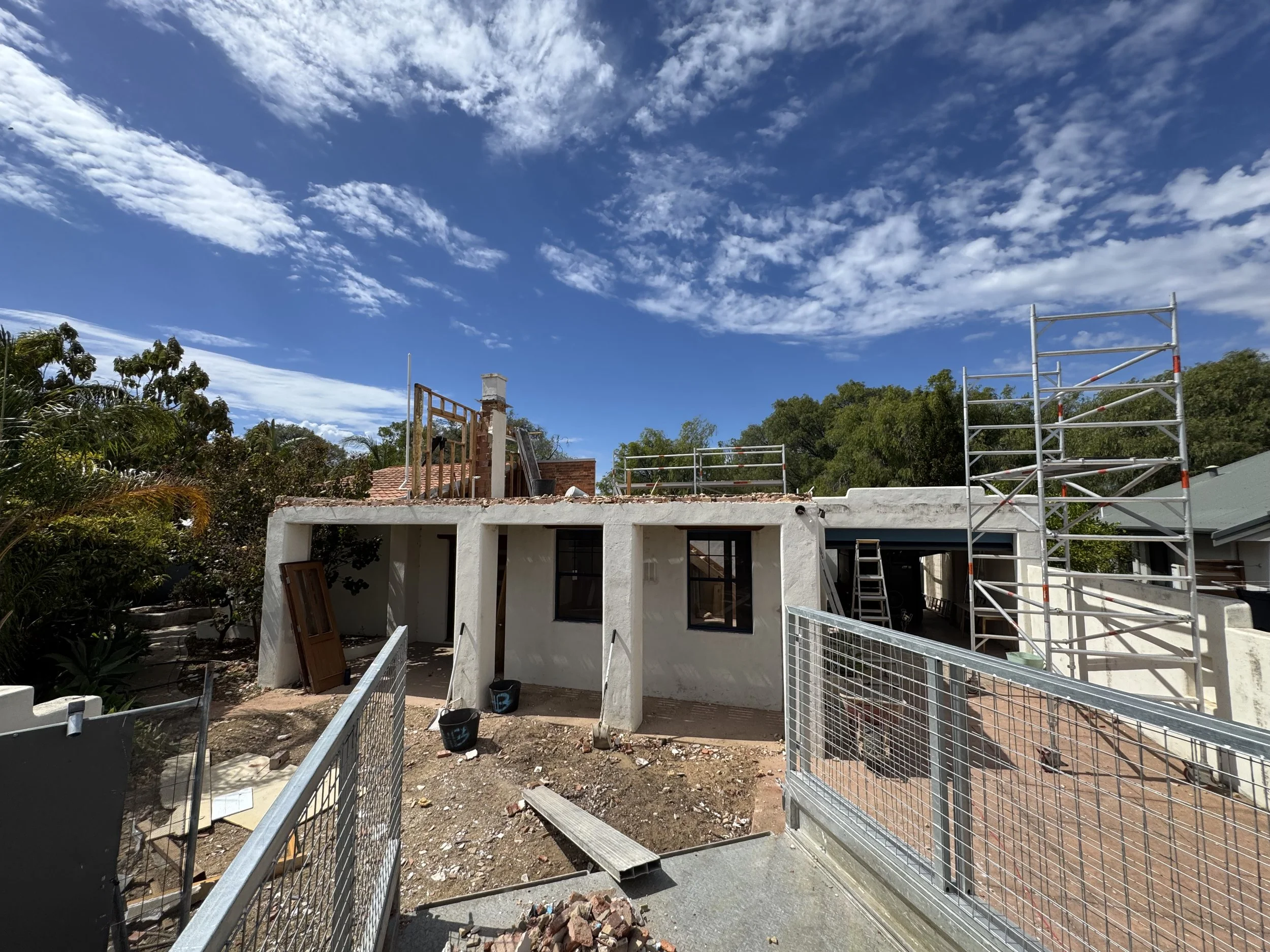 A house under construction with scaffolding, a partially finished roof, and construction materials around, set against a blue sky with scattered clouds.