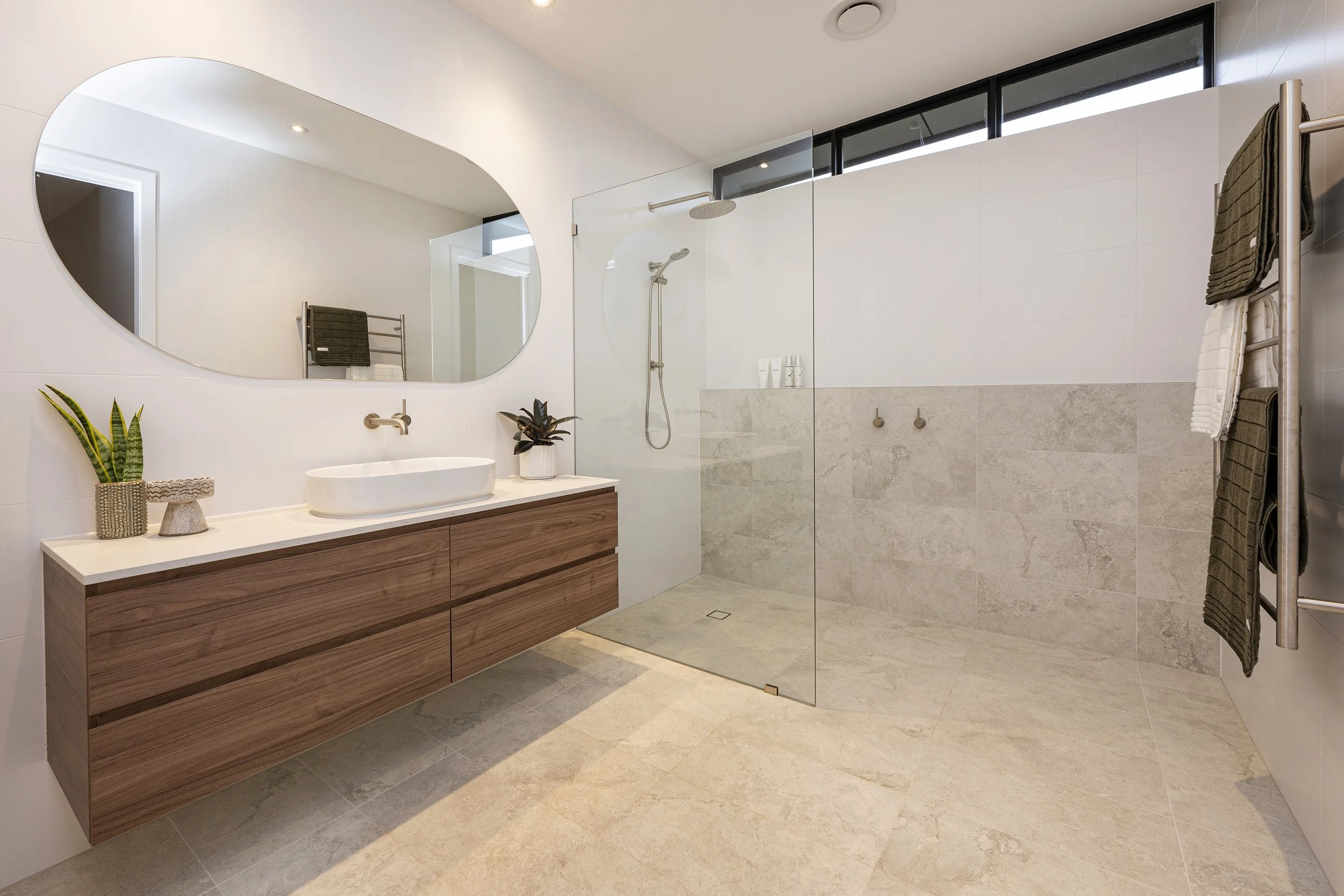 Modern bathroom with a floating wooden vanity, an oval mirror, a plant, and a walk-in shower with a glass partition and rain showerhead.