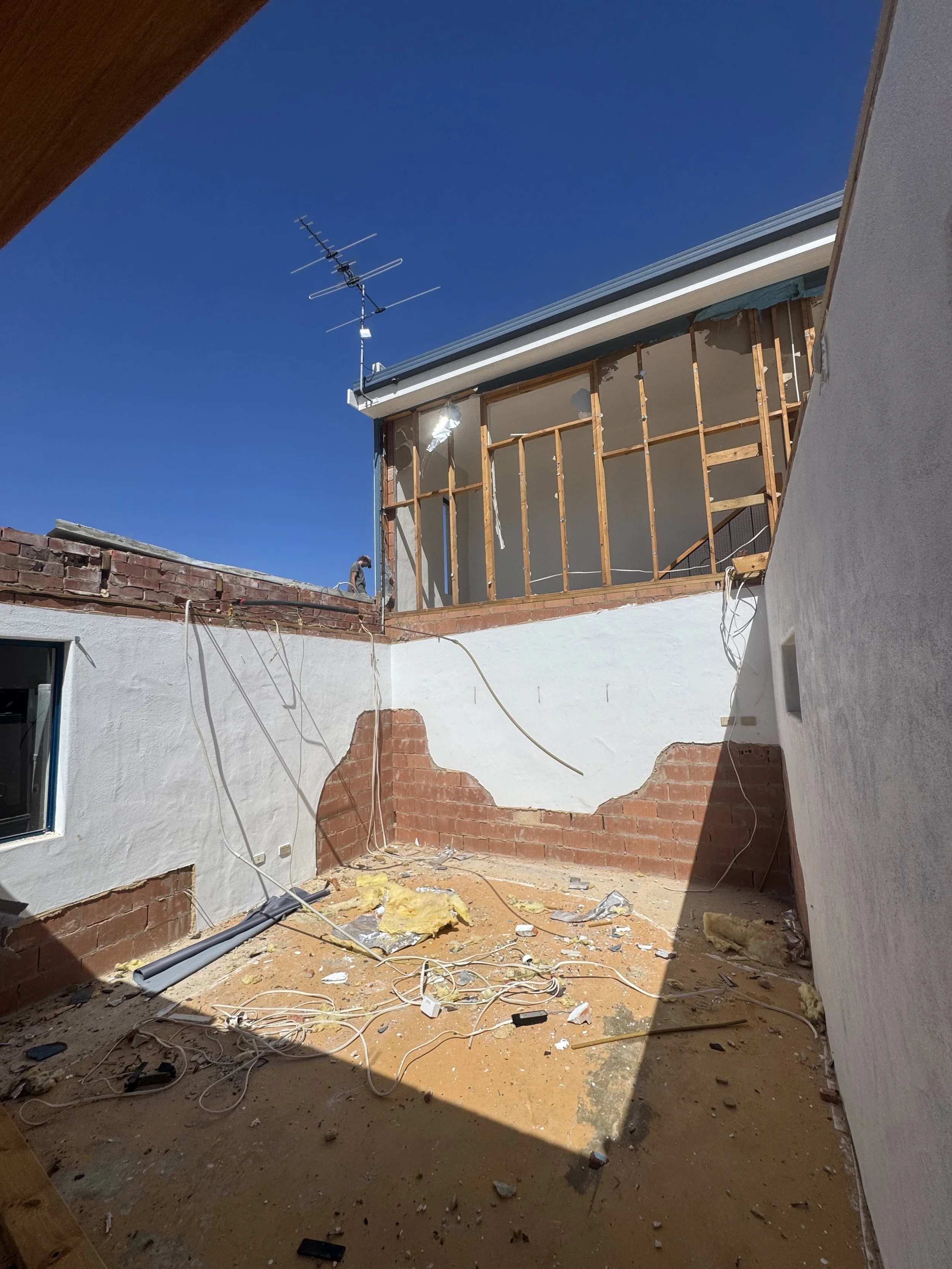 Construction site of a house undergoing renovation, with exposed wooden framing, construction debris, wires, and insulation, under a clear blue sky.