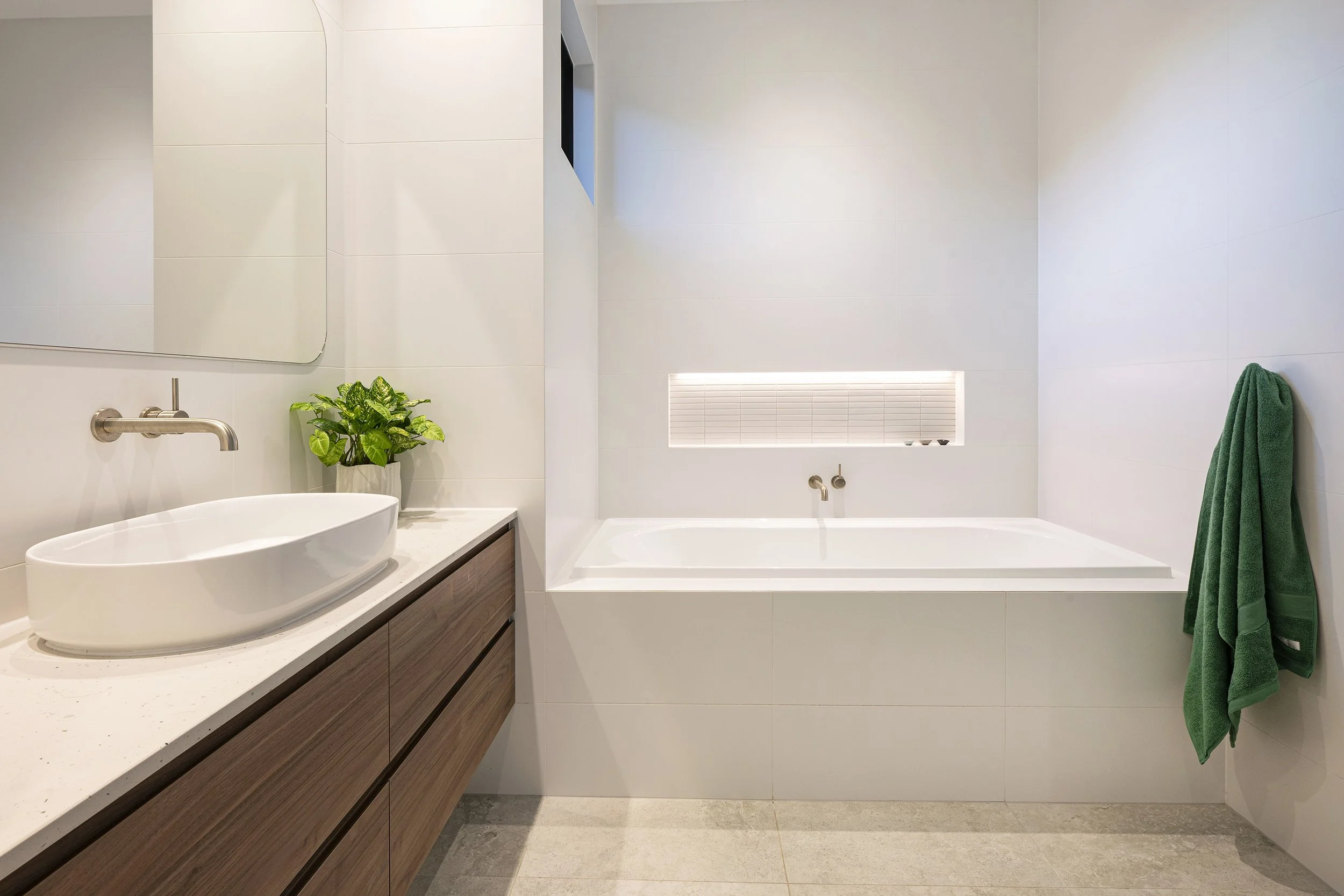 Modern bathroom with white tiled walls, a large mirror, a round vessel sink on a wooden vanity, a green towel hanging on a hook, a built-in shelf with lights, and a bathtub with a faucet.