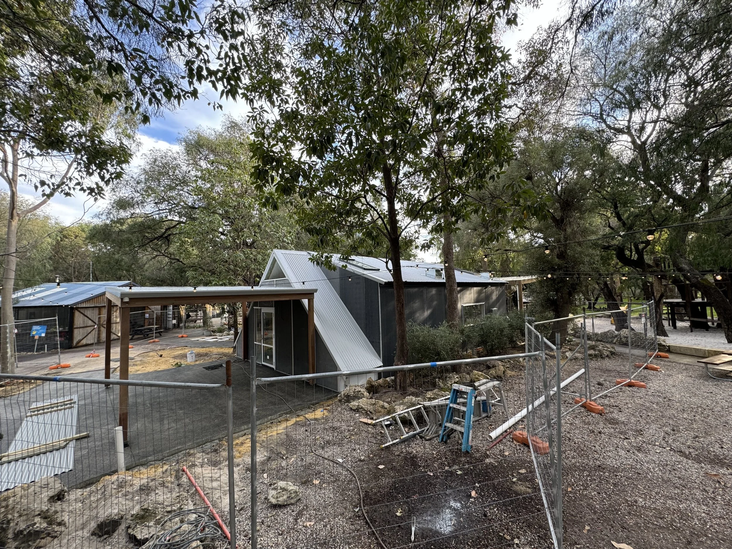 Construction site with fencing, trees, and a building with a metallic roof, surrounded by dirt and construction materials.