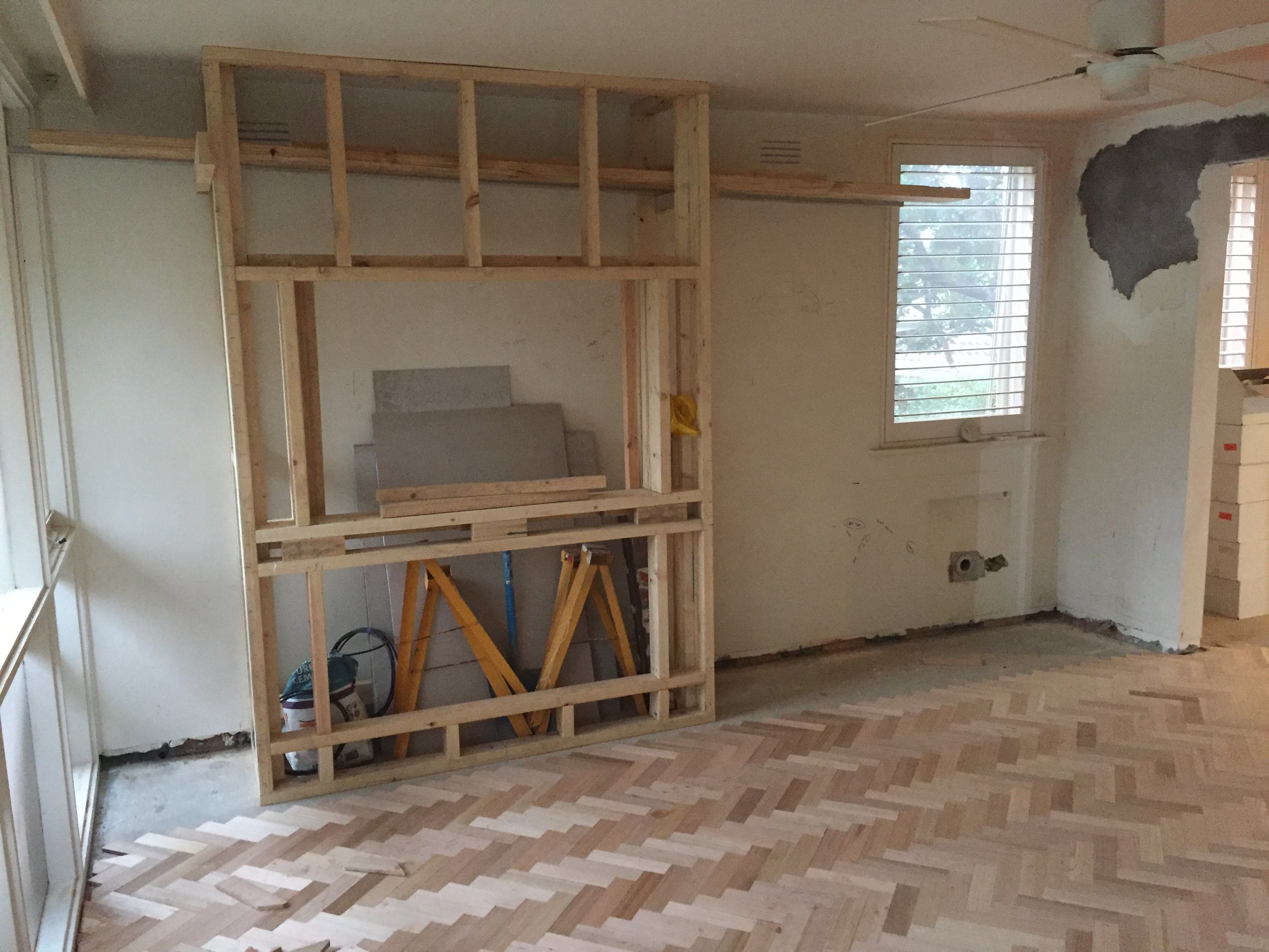 Interior of a room under renovation with a wooden framing structure, window with blinds, and no finished walls.