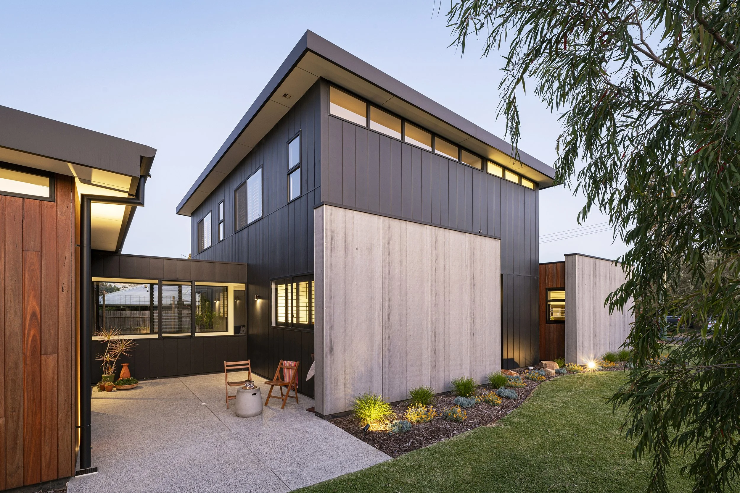 Modern two-story house with dark and light exterior panels, large windows, and a backyard patio with chairs and potted plants. There is landscaping with small shrubs and flower beds, and a tree on the right side. The house is illuminated by exterior 