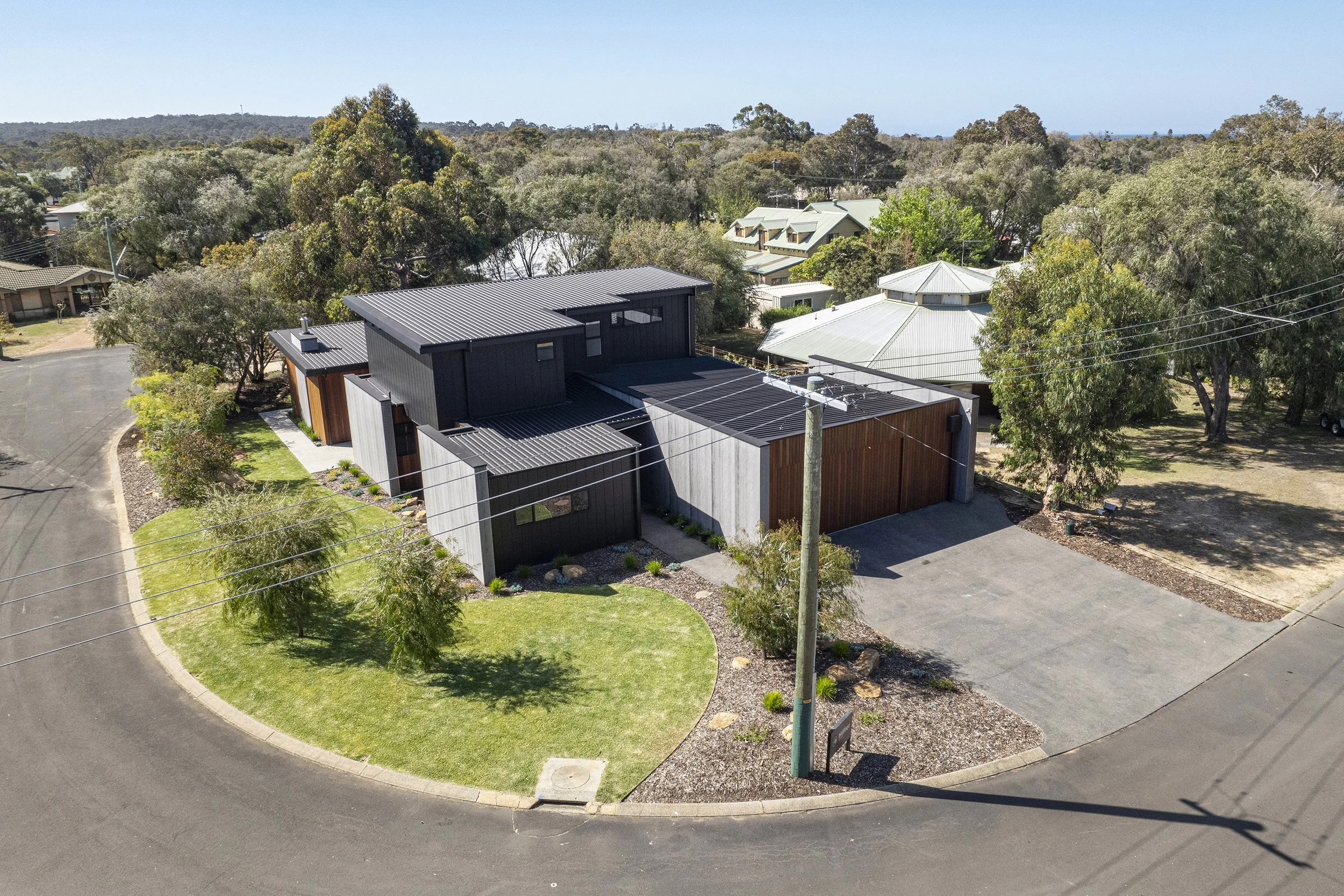 Aerial view of a modern house with black and wooden exterior, surrounded by trees and greenery, with a curved driveway and utility poles.