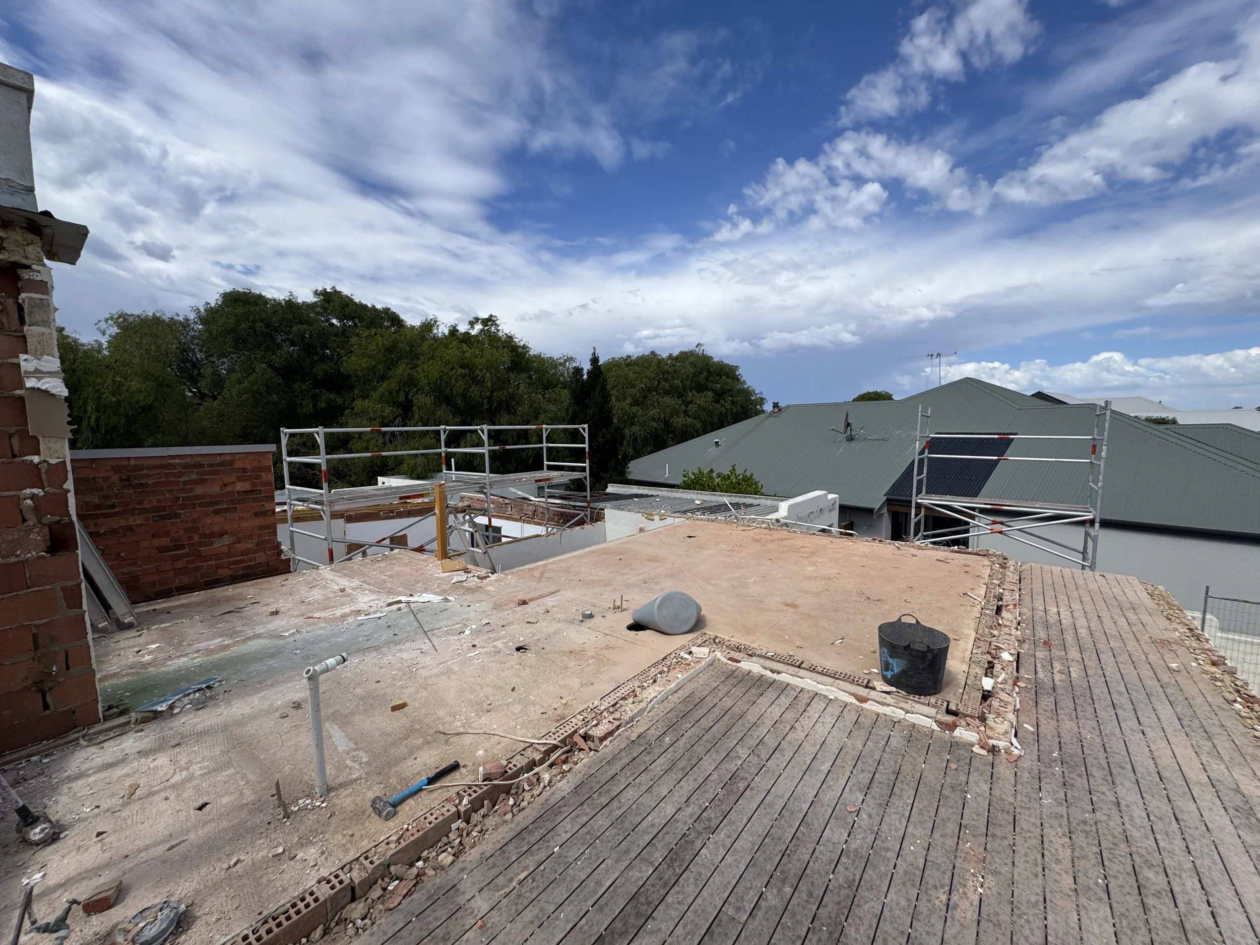 Construction site on a rooftop, with scaffolding, unfinished brick walls, and a view of roofs and trees under a blue sky with clouds.