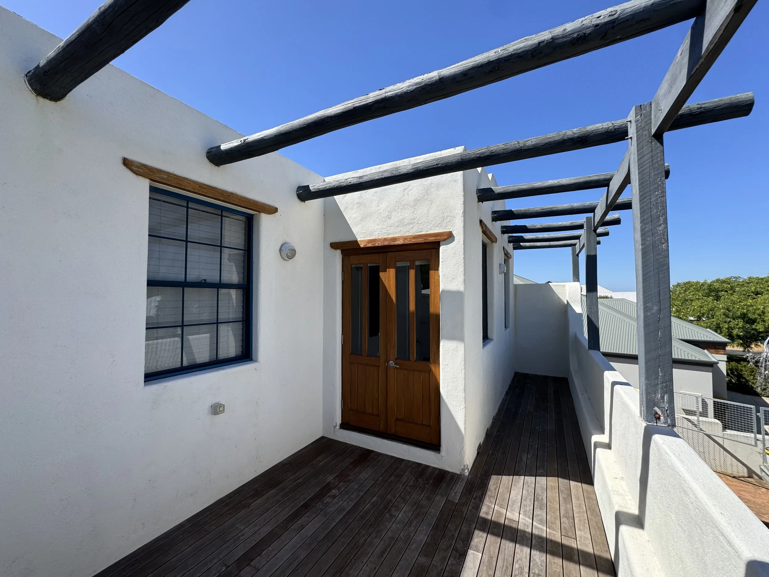 Residential balcony with dark wooden decking, a white stucco wall, and a wooden door with glass panels. There are wooden beams extending outward from the building structure, and a window with black grid frames on the wall. The sky is clear and blue, 