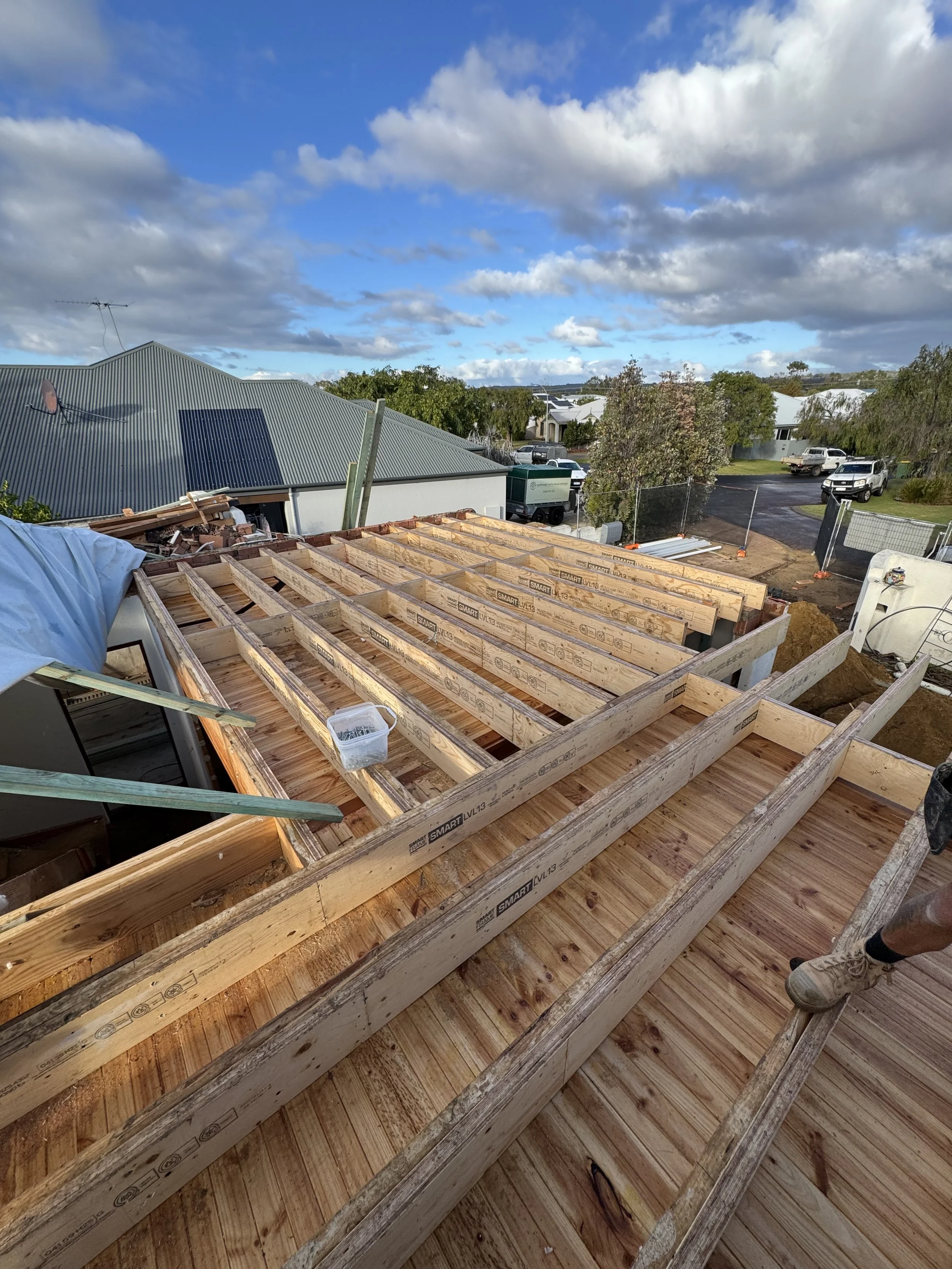 View of a construction site with wooden framing for a building, a partly cloudy sky, nearby trees, and parked vehicles in the background.