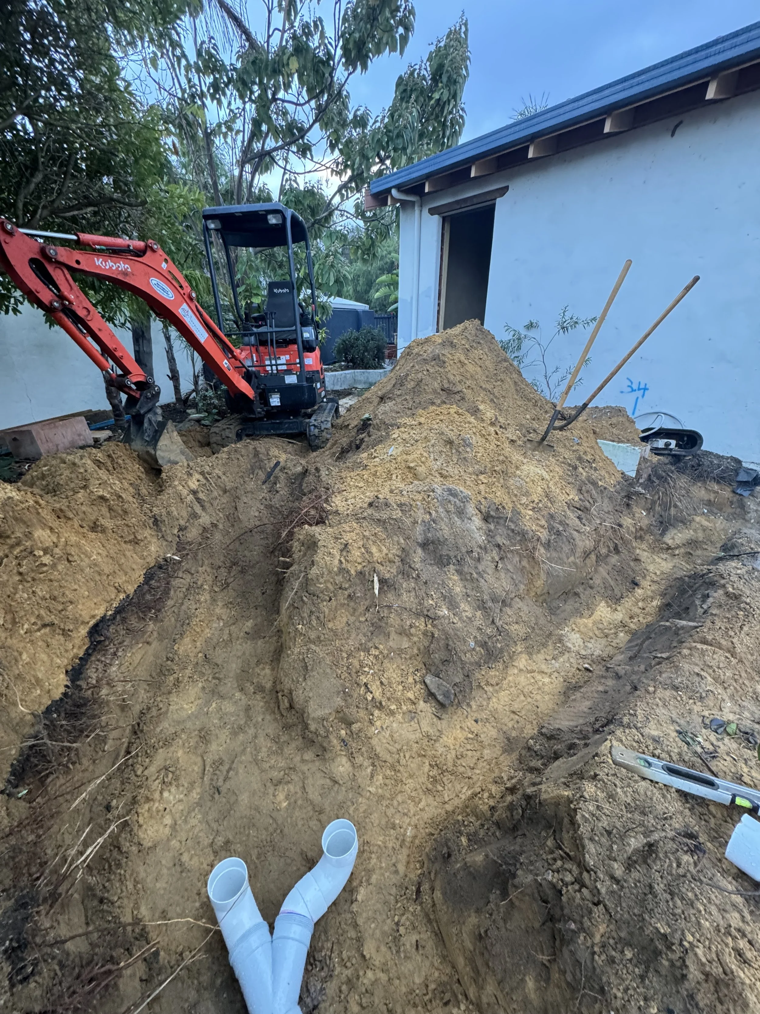 Construction site with a small excavator and PVC pipes in a dirt trench next to a house with a white exterior wall.