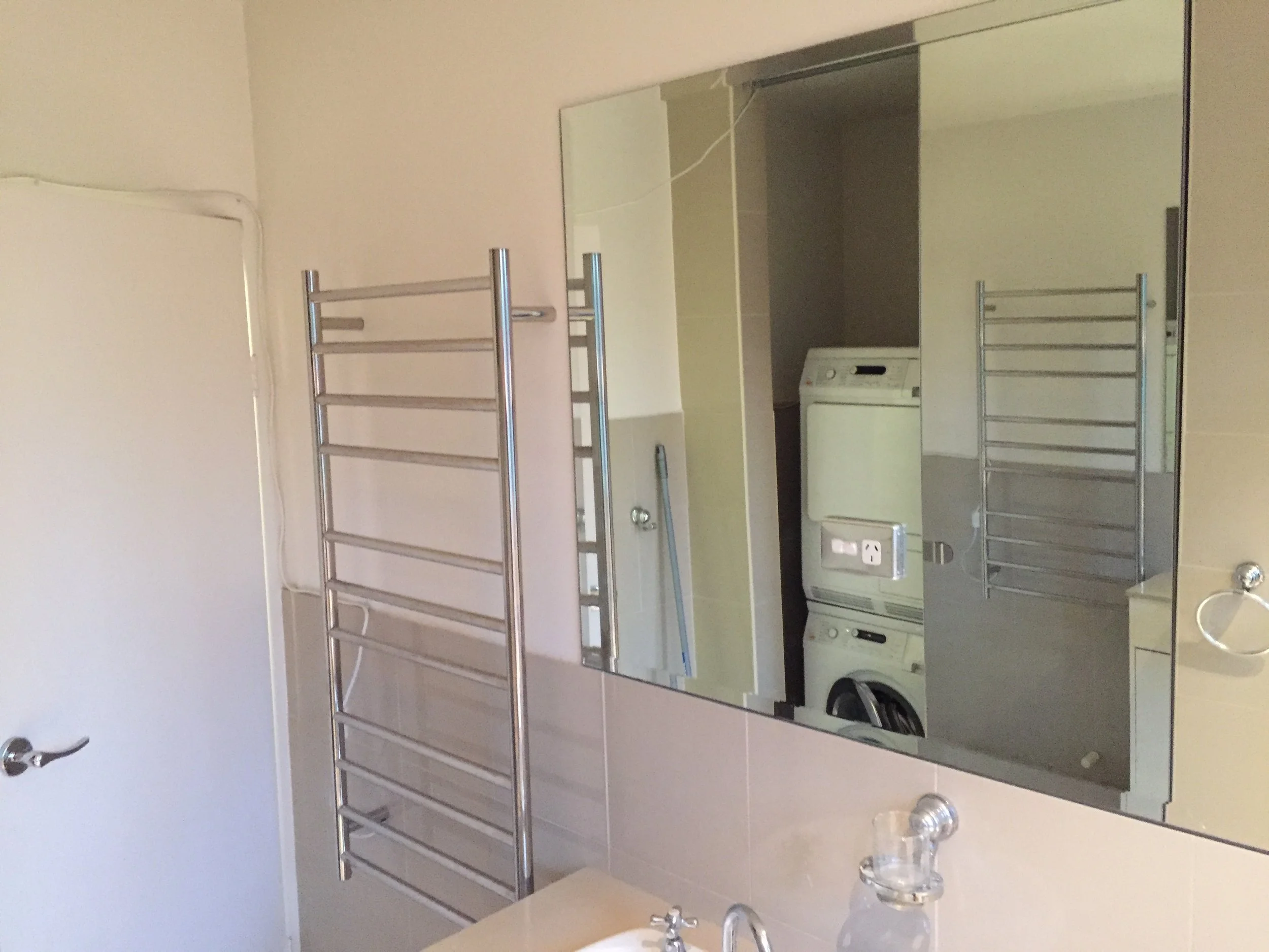 A bathroom mirror with a metal towel rack and a visible washing machine underneath. The wall is light-colored, and part of a closed door with a metallic doorknob is visible on the left.