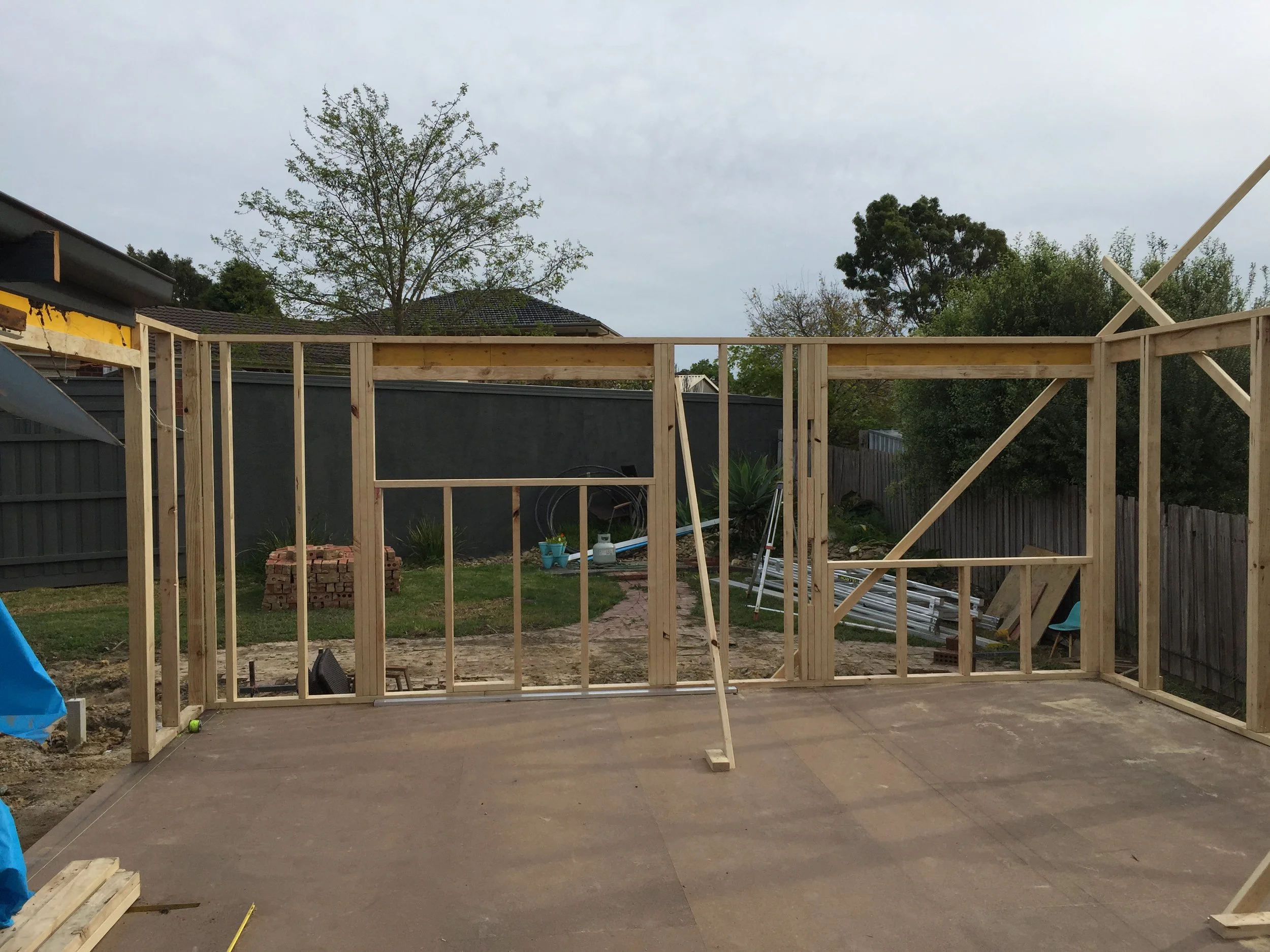 Wooden framing for a building under construction in a backyard with trees and a gray fence, some construction materials and tools visible.