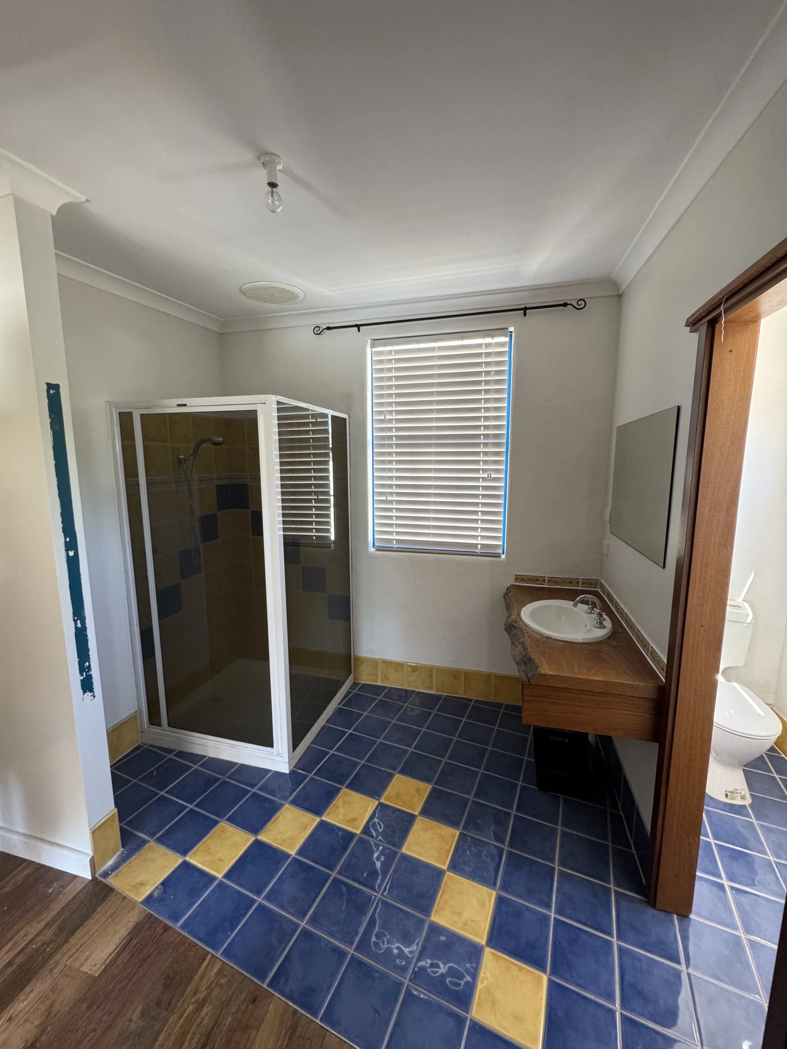 A bathroom with a blue and yellow tiled floor, a shower stall in the corner, a small sink with a stone countertop, and a window with horizontal blinds.