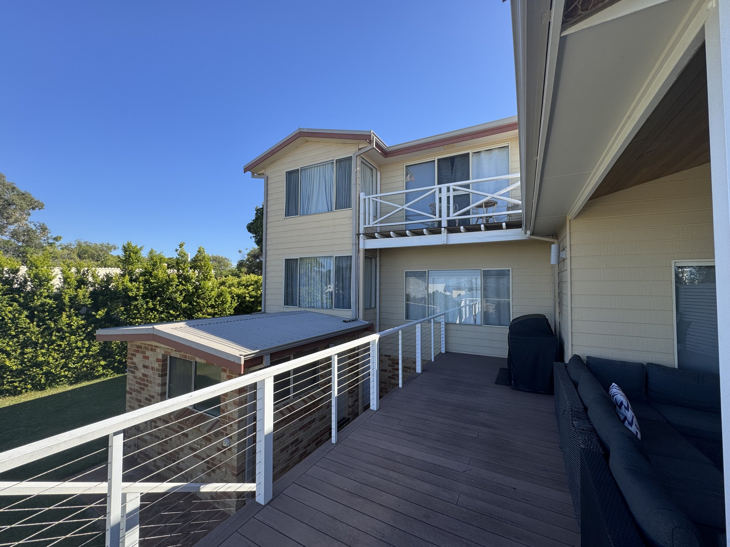 Balcony view of a beige two-story house with large windows and a small white railing patio, surrounded by green trees under a clear blue sky during daytime.