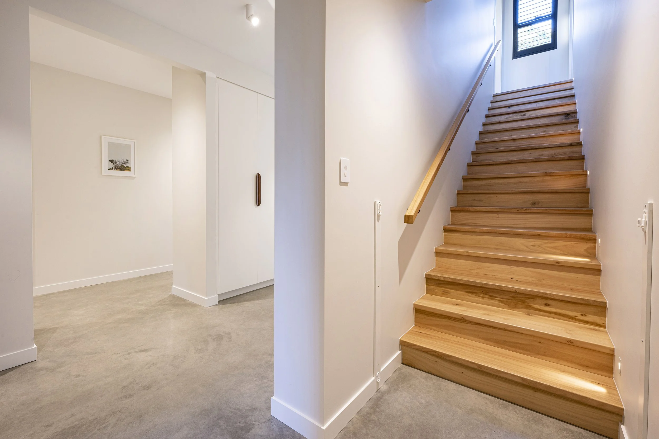 Interior view of a house with a wooden staircase, white walls, a small framed picture on the wall, and a window at the top of the stairs letting in natural light.