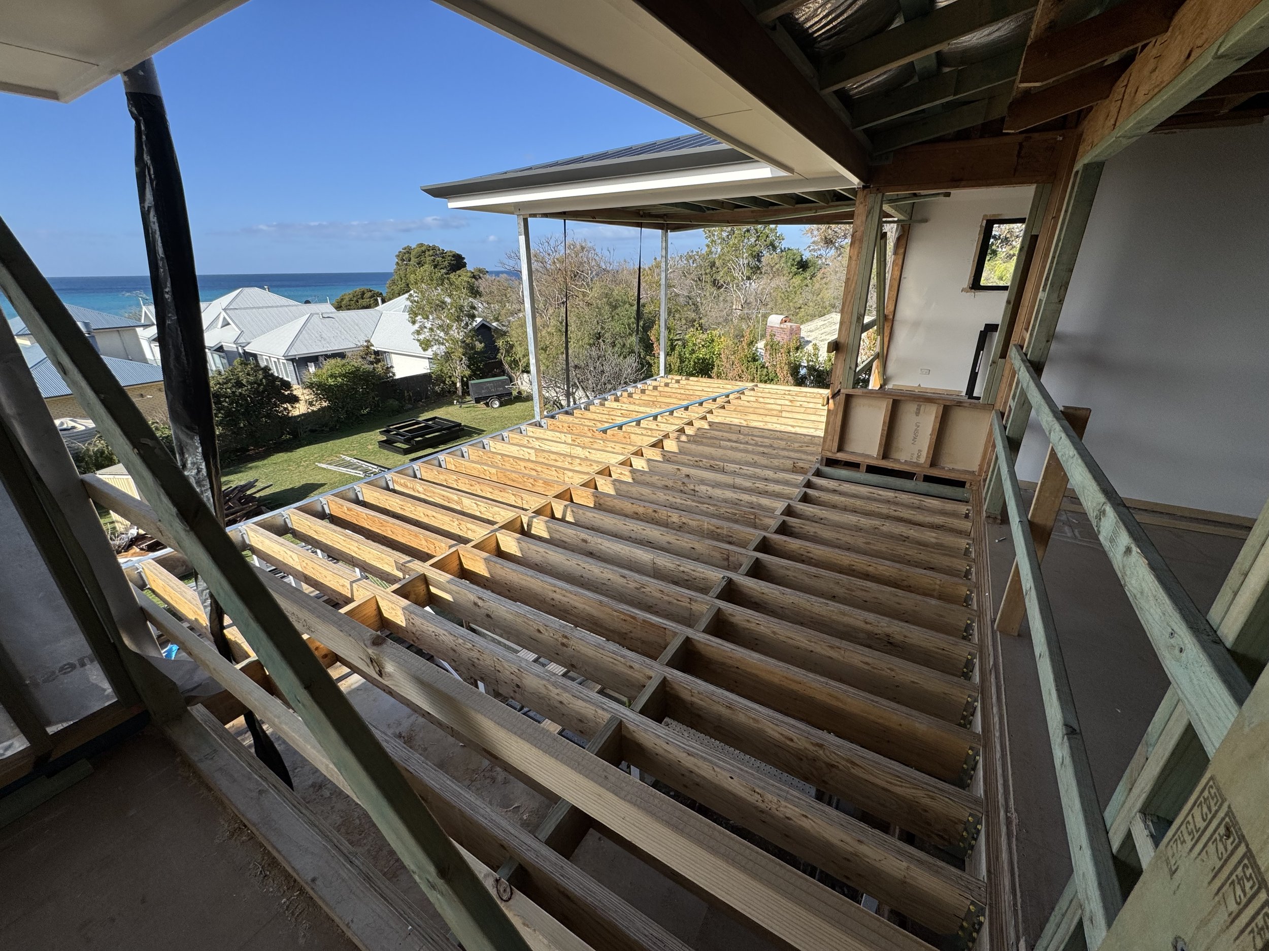 Under construction building with wooden beams on a residential area overlooking rooftops, trees, and the ocean in the distance.
