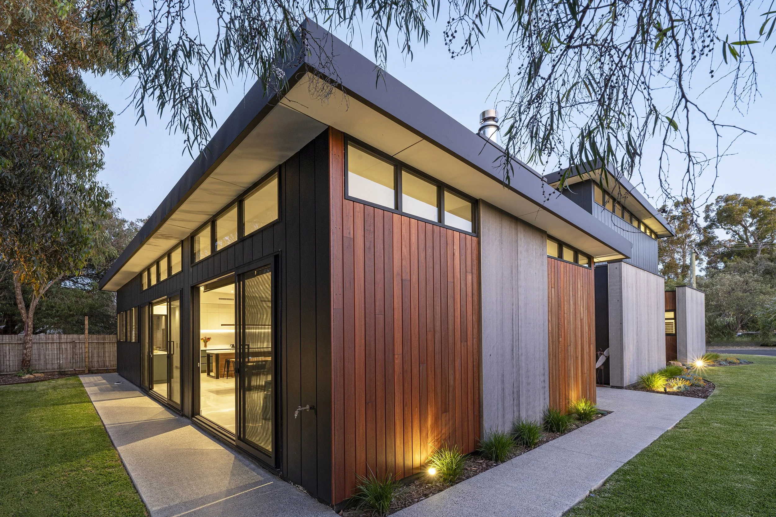 Modern two-story house with large glass windows, wooden and concrete exterior siding, surrounded by a manicured lawn, trees, and exterior lighting at dusk.
