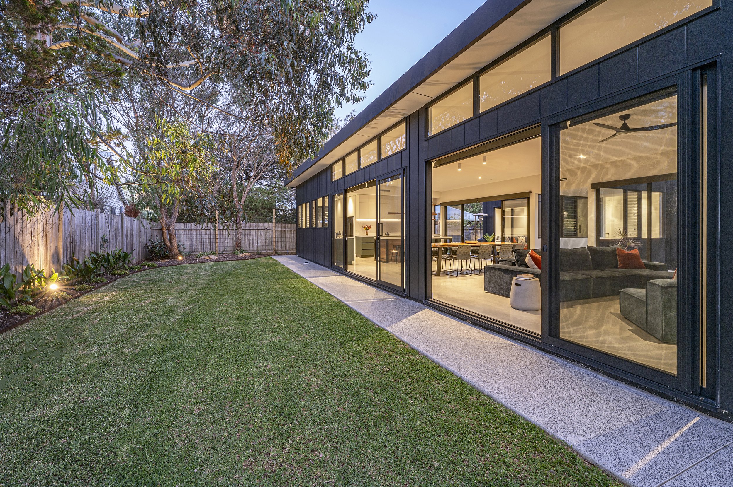 Modern house with large glass windows and sliding doors, showcasing the interior living room and dining area, with a well-maintained backyard, trees, and outdoor lighting at dusk.