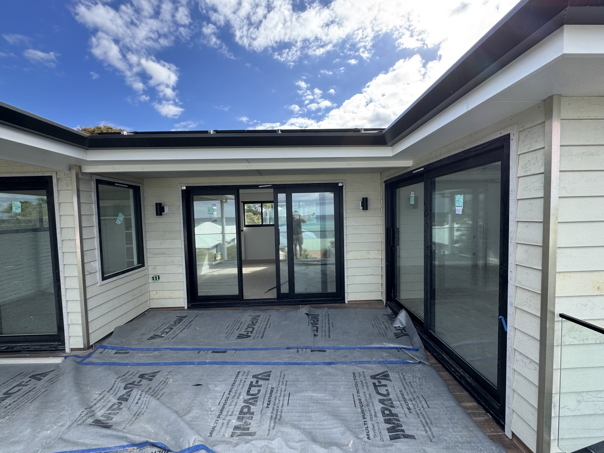 View of a house undergoing construction, with large glass sliding doors and windows, and protective covering on the floor, under a partly cloudy sky.